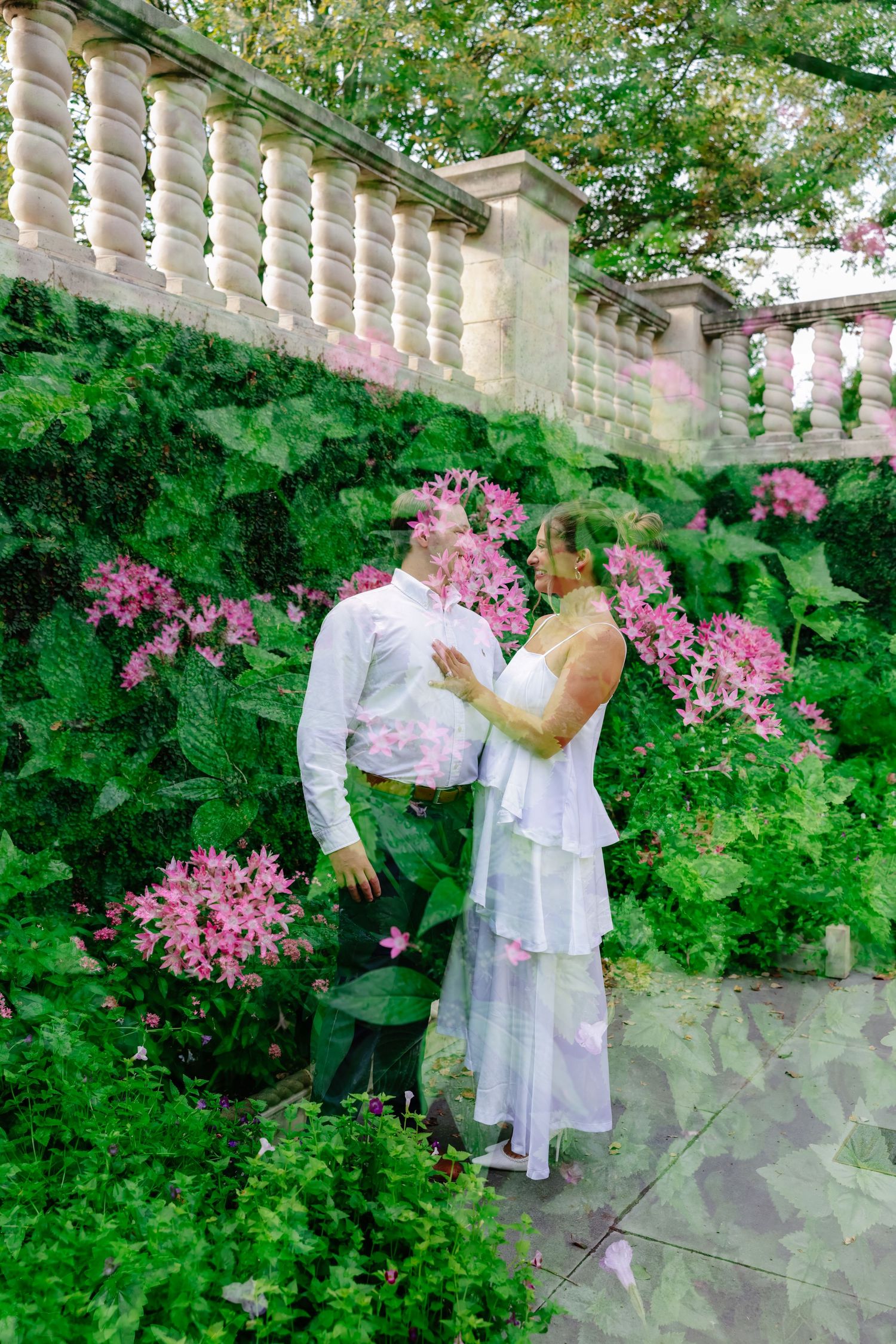 Couple in white attire embracing among lush green ferns and pink flowers beside a stone balustrade.