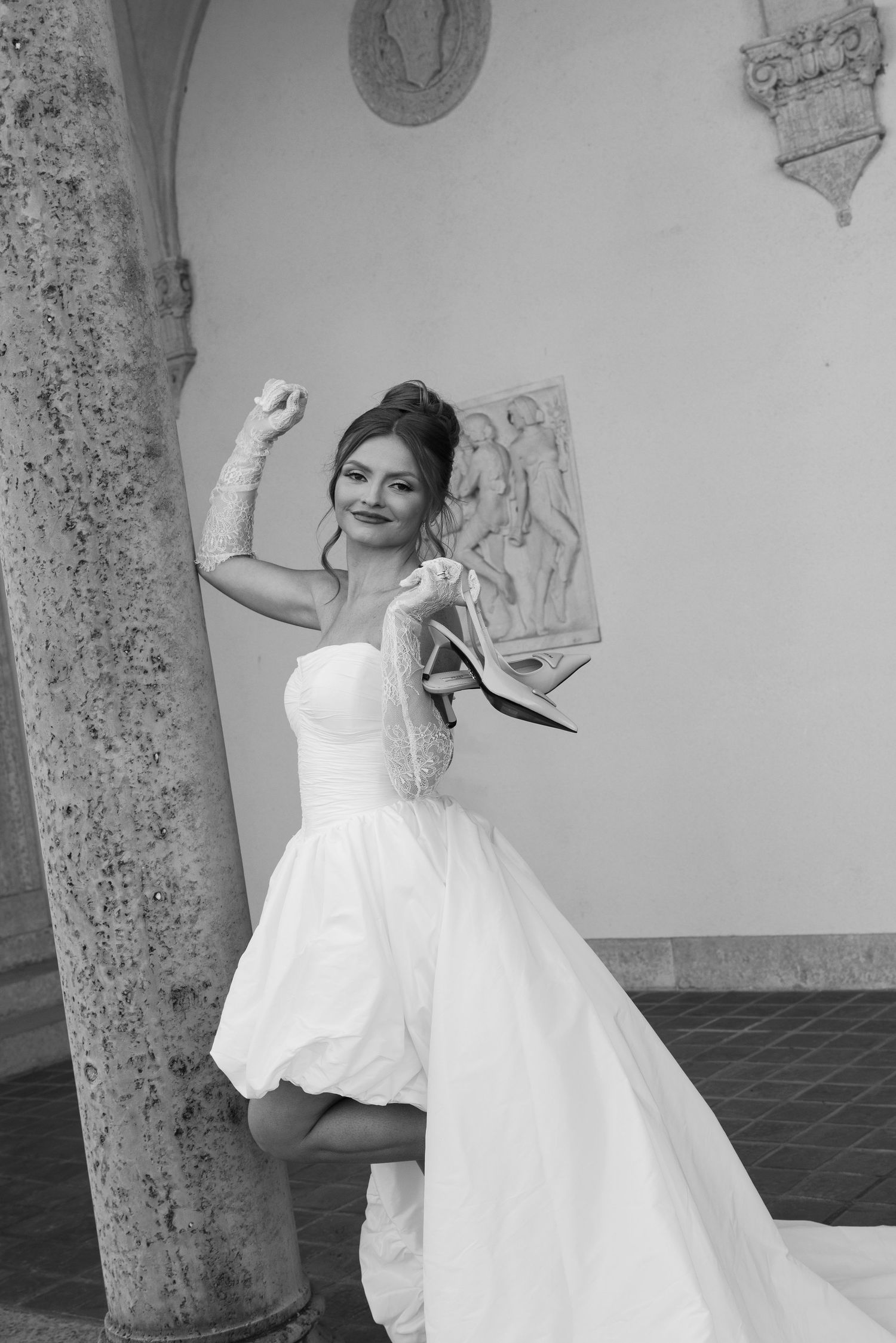 A joyful bride in a white gown poses by a stone column in a black and white photo.