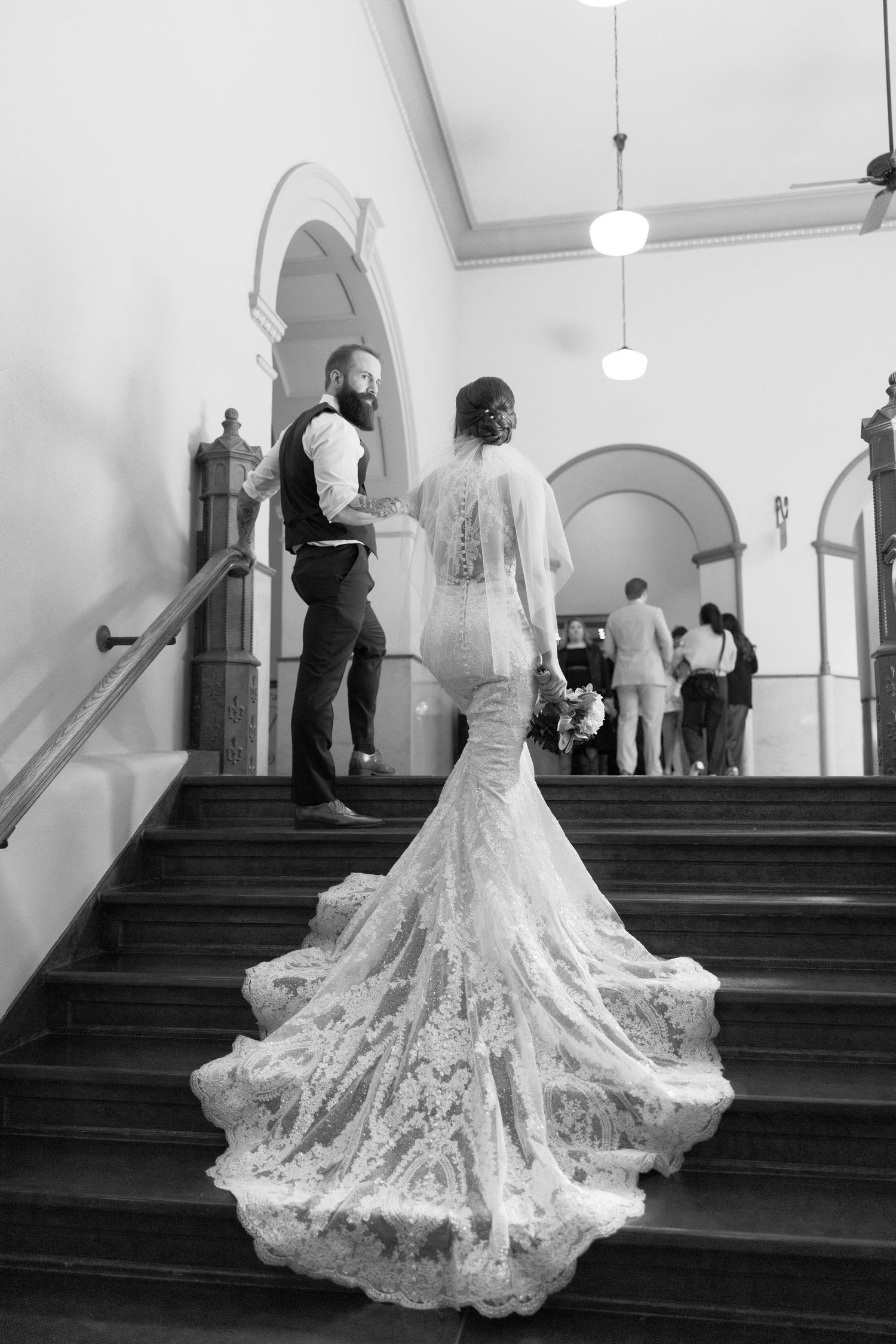Black and white photo of bride in lace gown with long train ascending stairs with groom at elegant wedding venue.