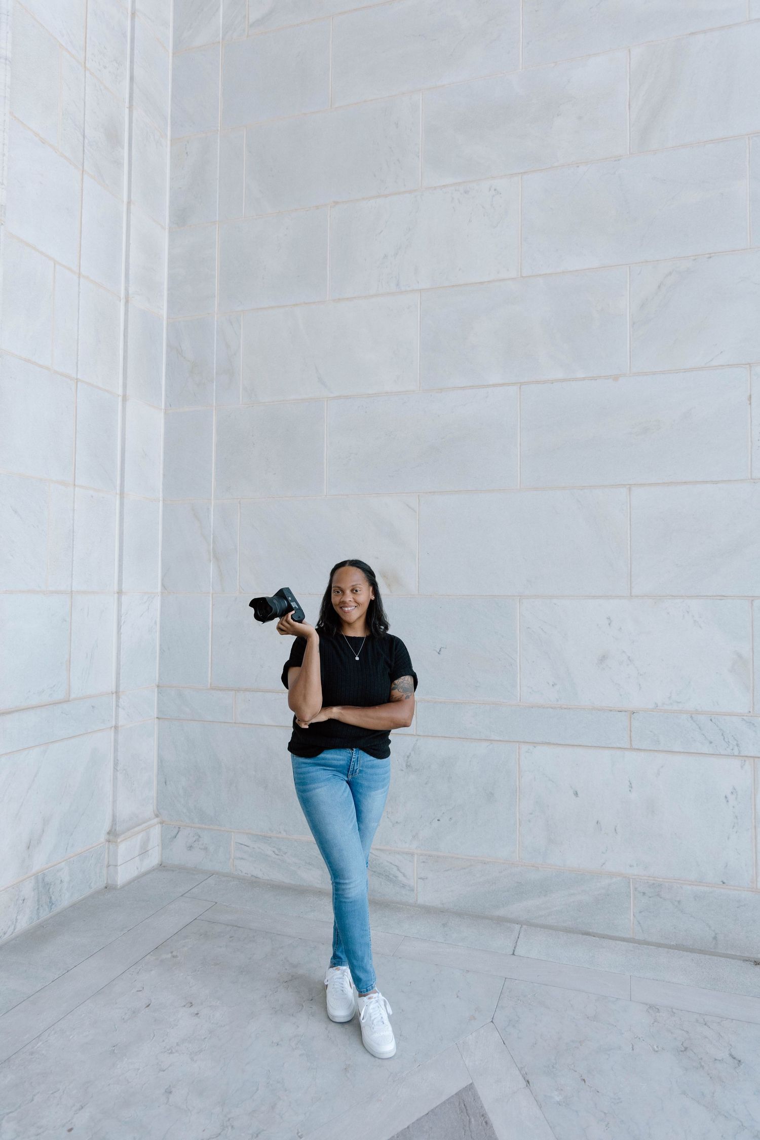 A photographer in jeans and white sneakers poses with a camera in different positions against a white marble wall.