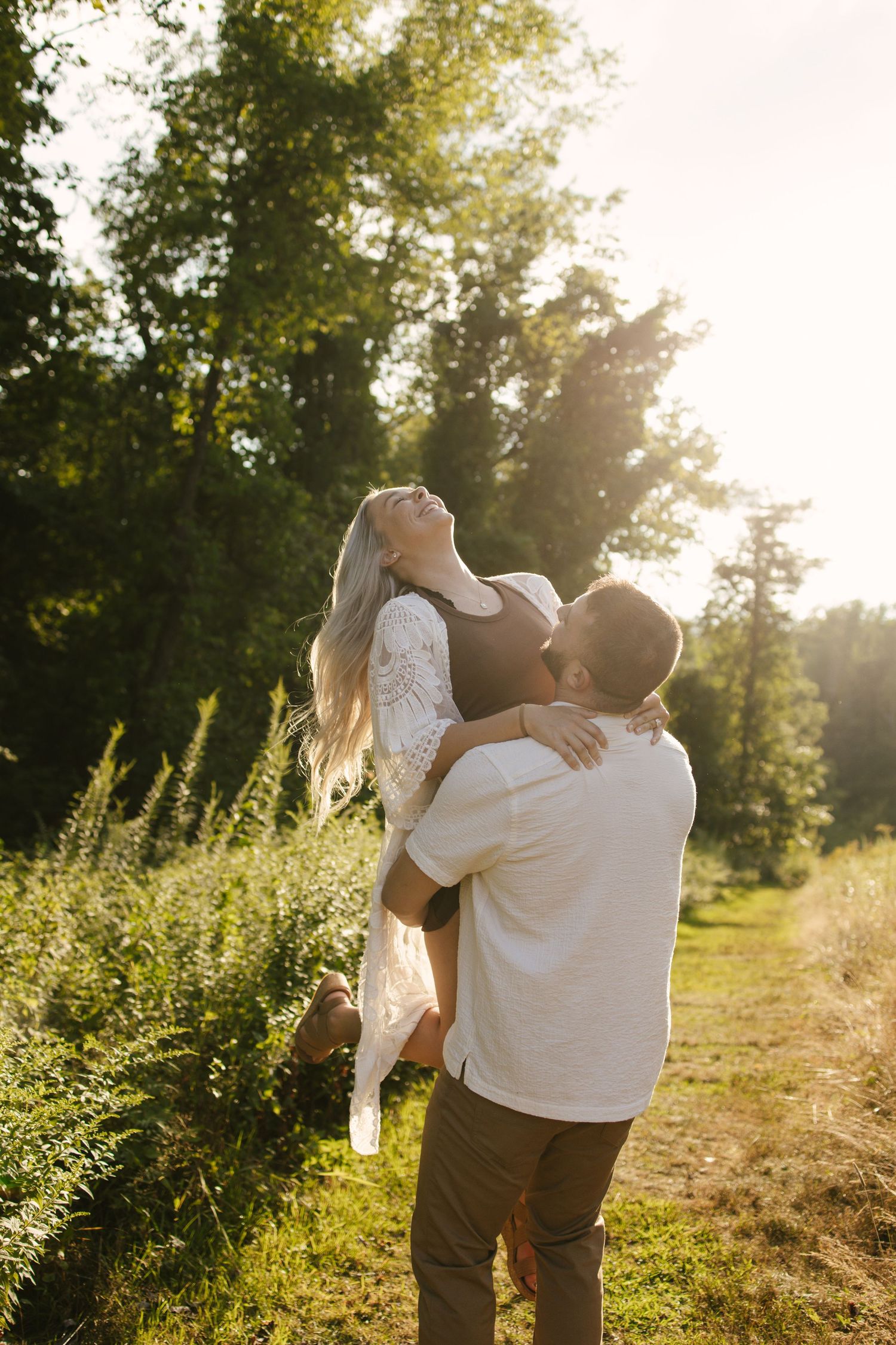 A couple embraces in a sunlit field with golden light streaming through trees during a romantic outdoor photoshoot.