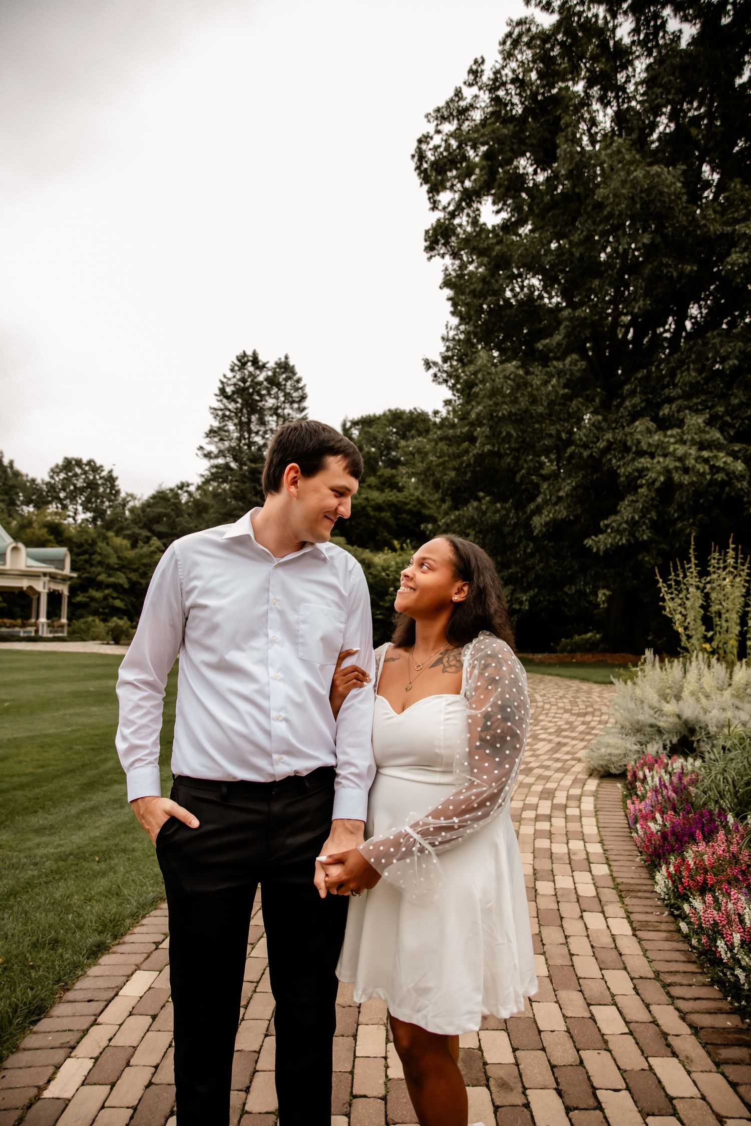 A couple in wedding attire walks together along a stone pathway bordered by colorful flowers and greenery.