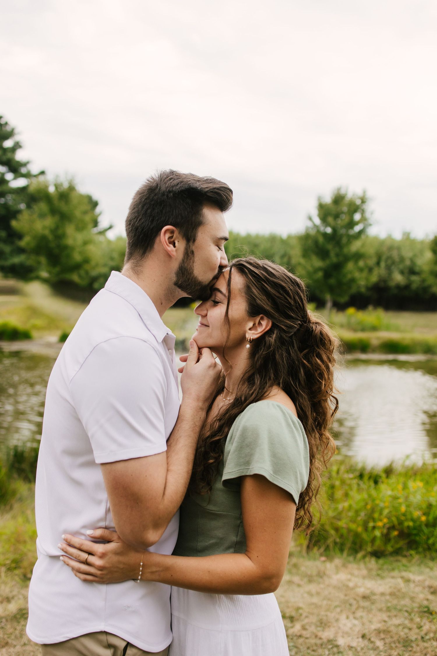 A romantic couple shares intimate kisses by a serene lake surrounded by lush greenery on a summer day.