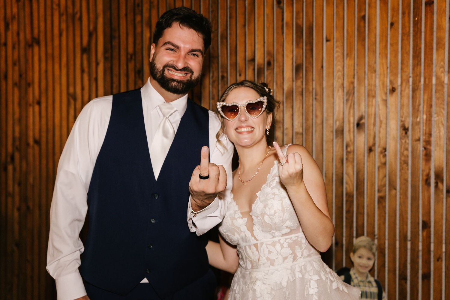 Joyful couple in wedding attire share laughter and smiles in front of rustic wooden wall backdrop.