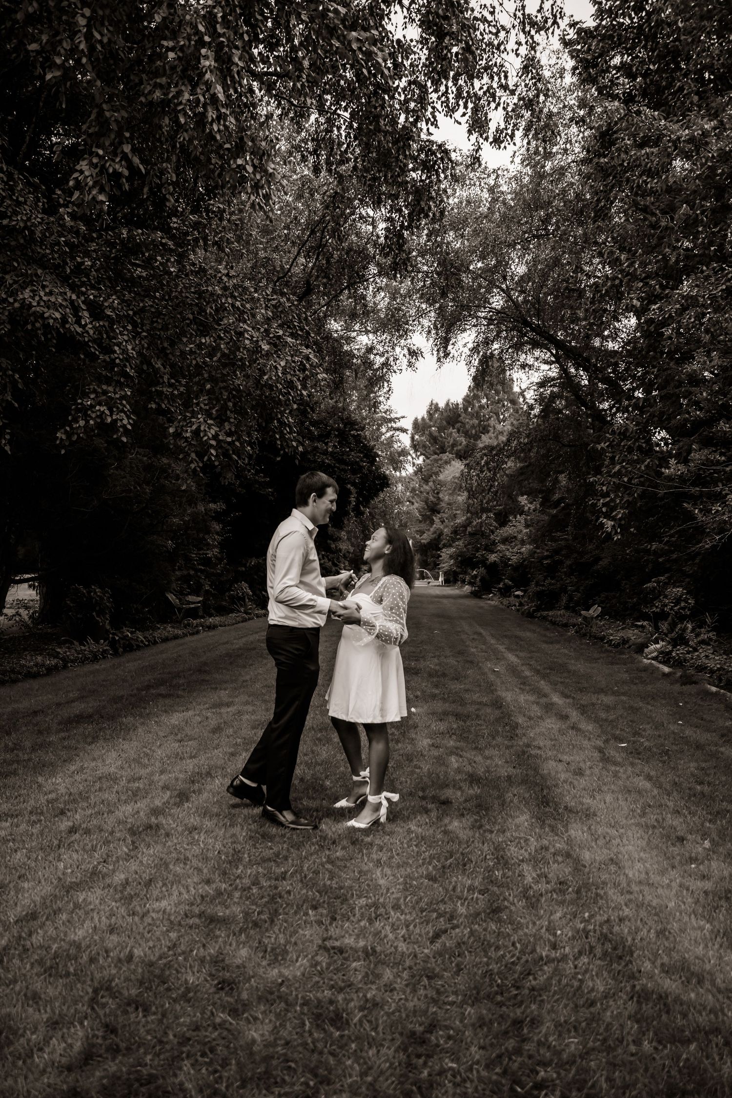 A romantic black and white photo series showing a couple standing together on a tree-lined path in a park setting.
