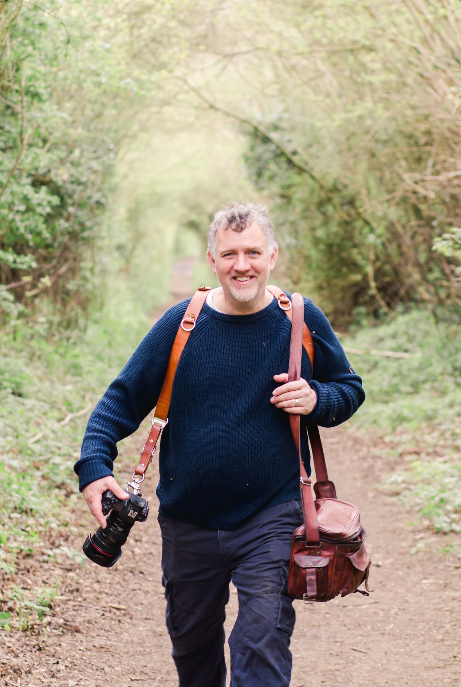 Photographer carrying gear walks along wooded trail with cheerful expression.