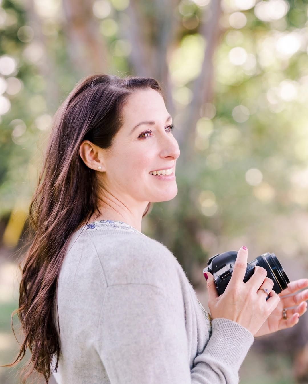 Photographer with long dark hair holding camera outdoors in natural setting.