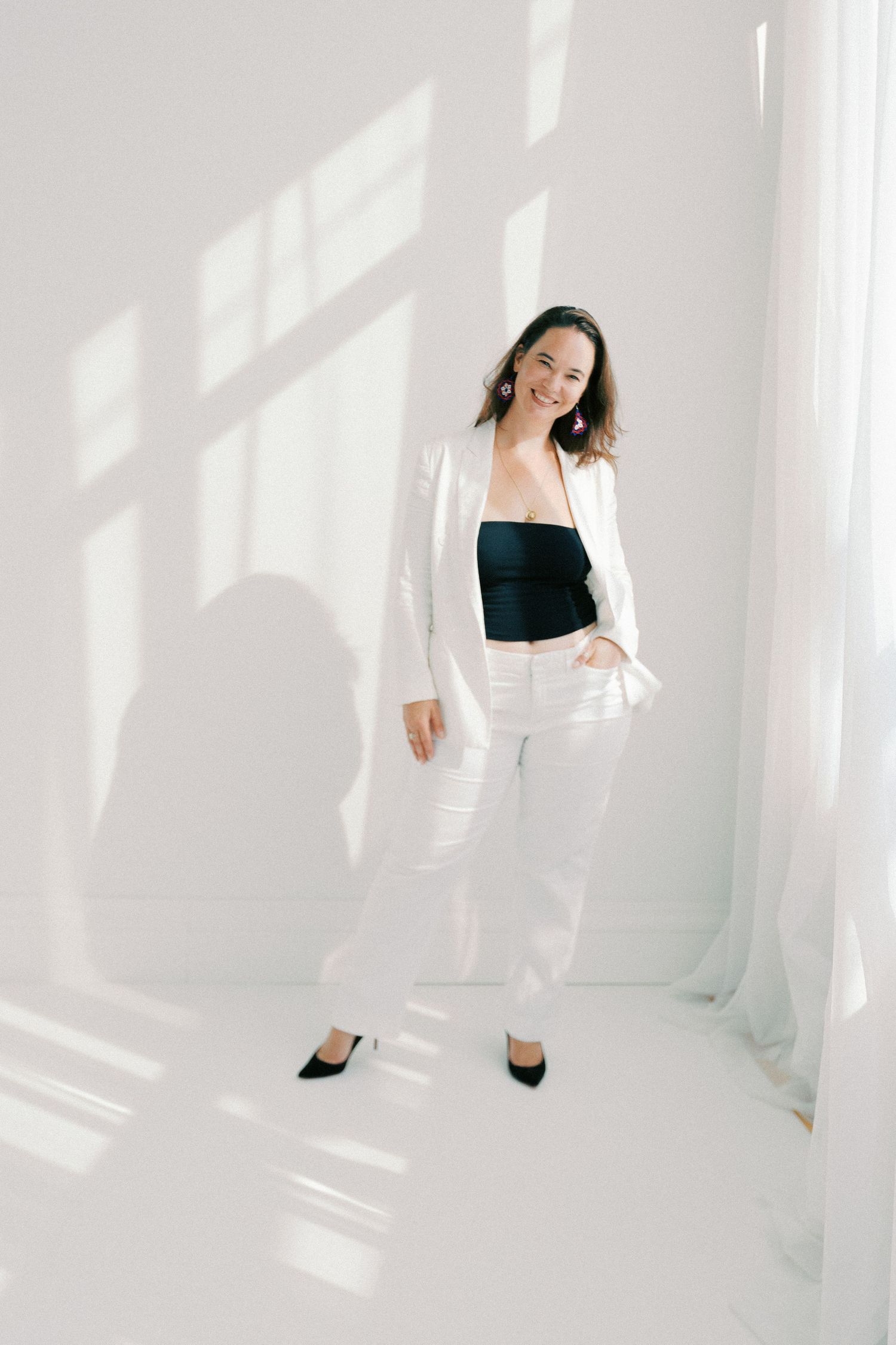 Woman in white blazer and pants with black crop top posing confidently in bright white studio setting.
