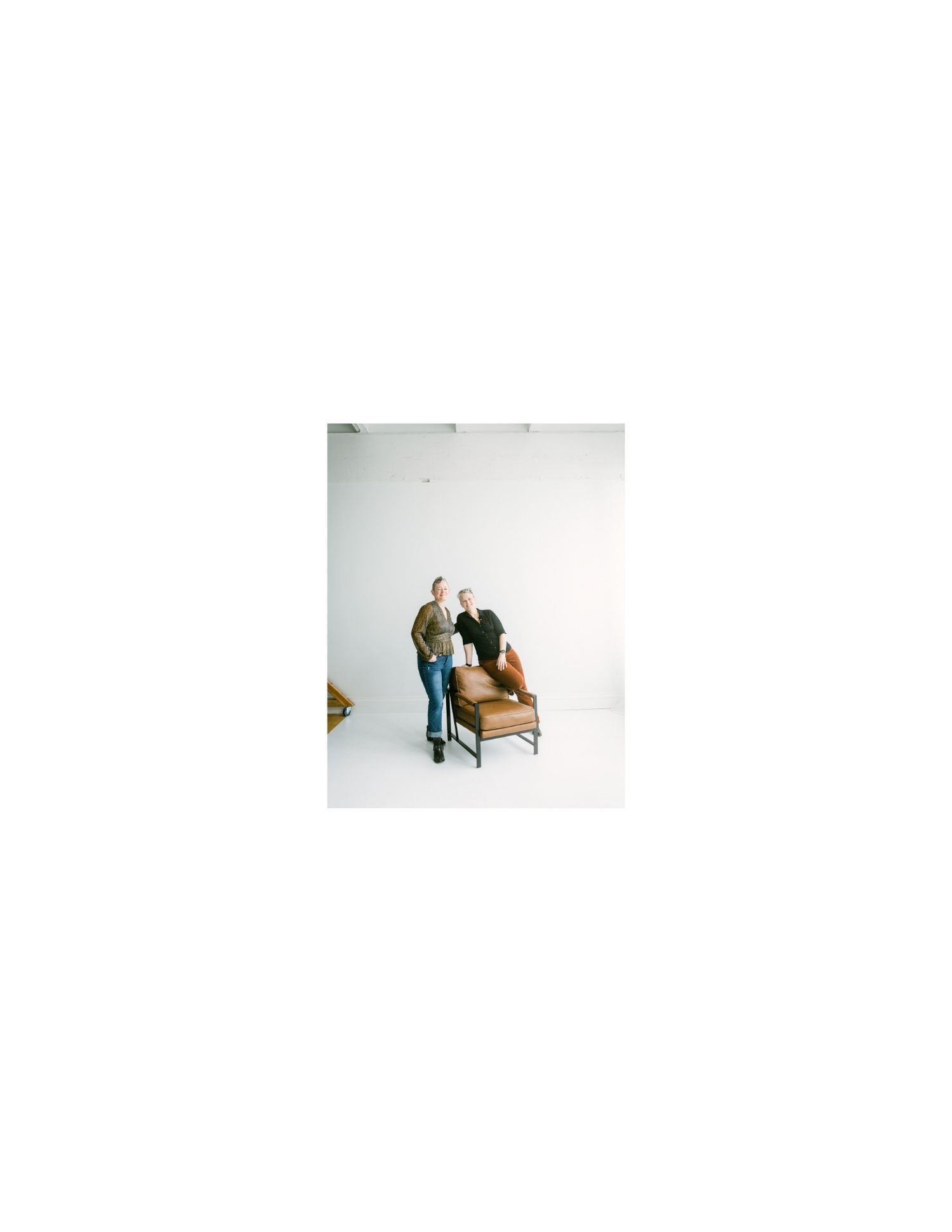 Two women stand and sit around a brown leather chair in a bright white photography studio.