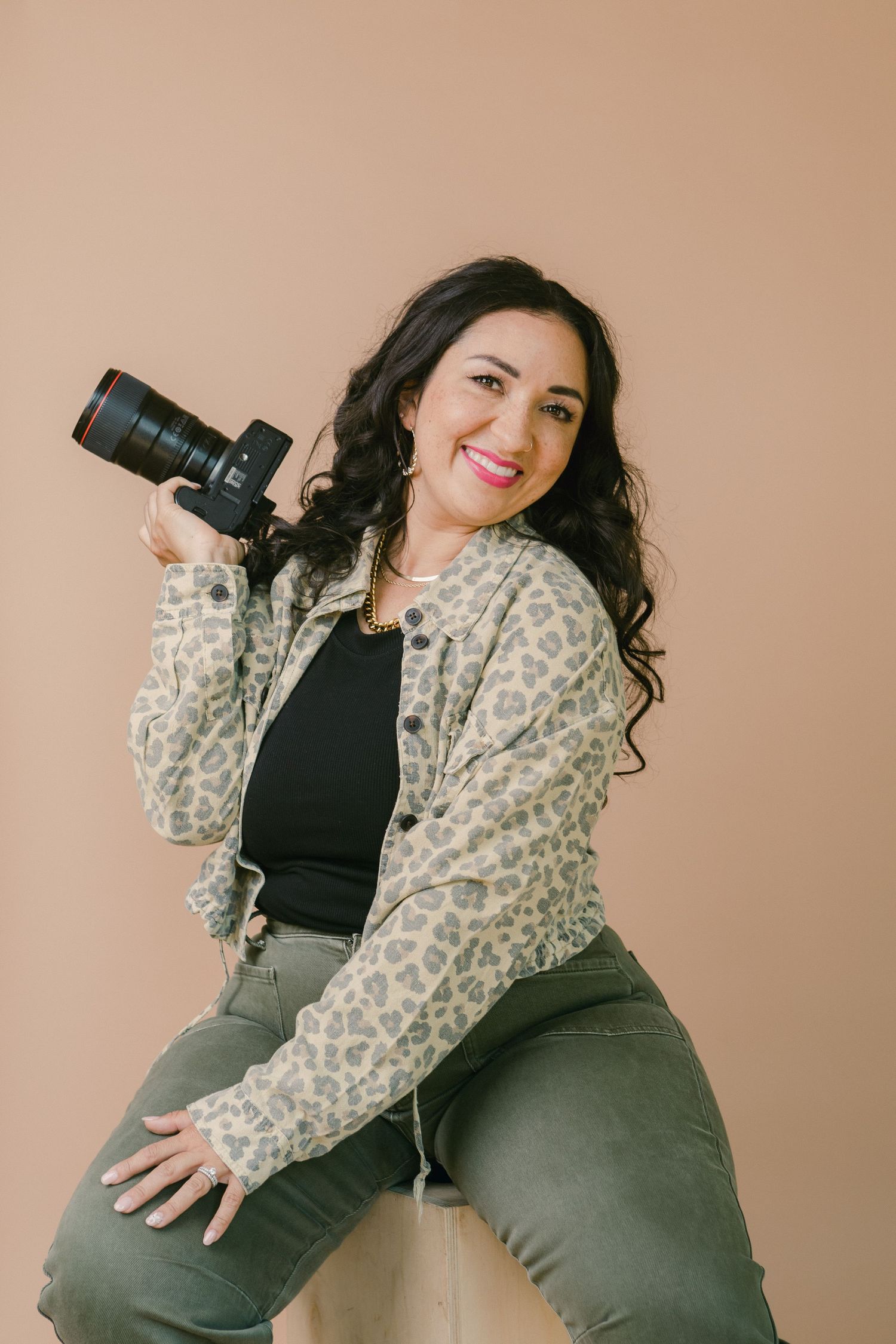 Woman in leopard print jacket holding a DSLR camera, smiling while seated against a beige background.