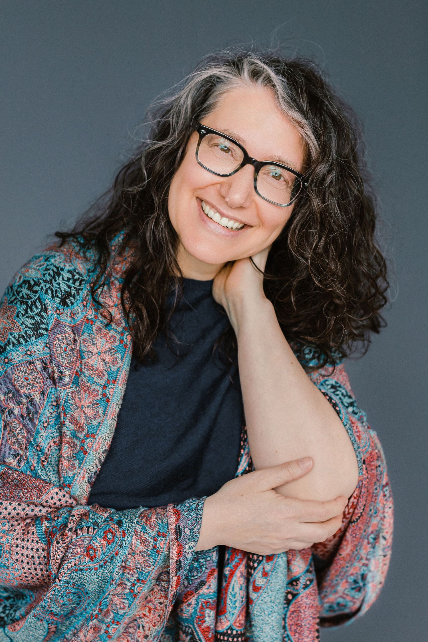 Smiling middle-aged woman with curly gray-streaked hair, black glasses, and colorful kimono poses against gray background.
