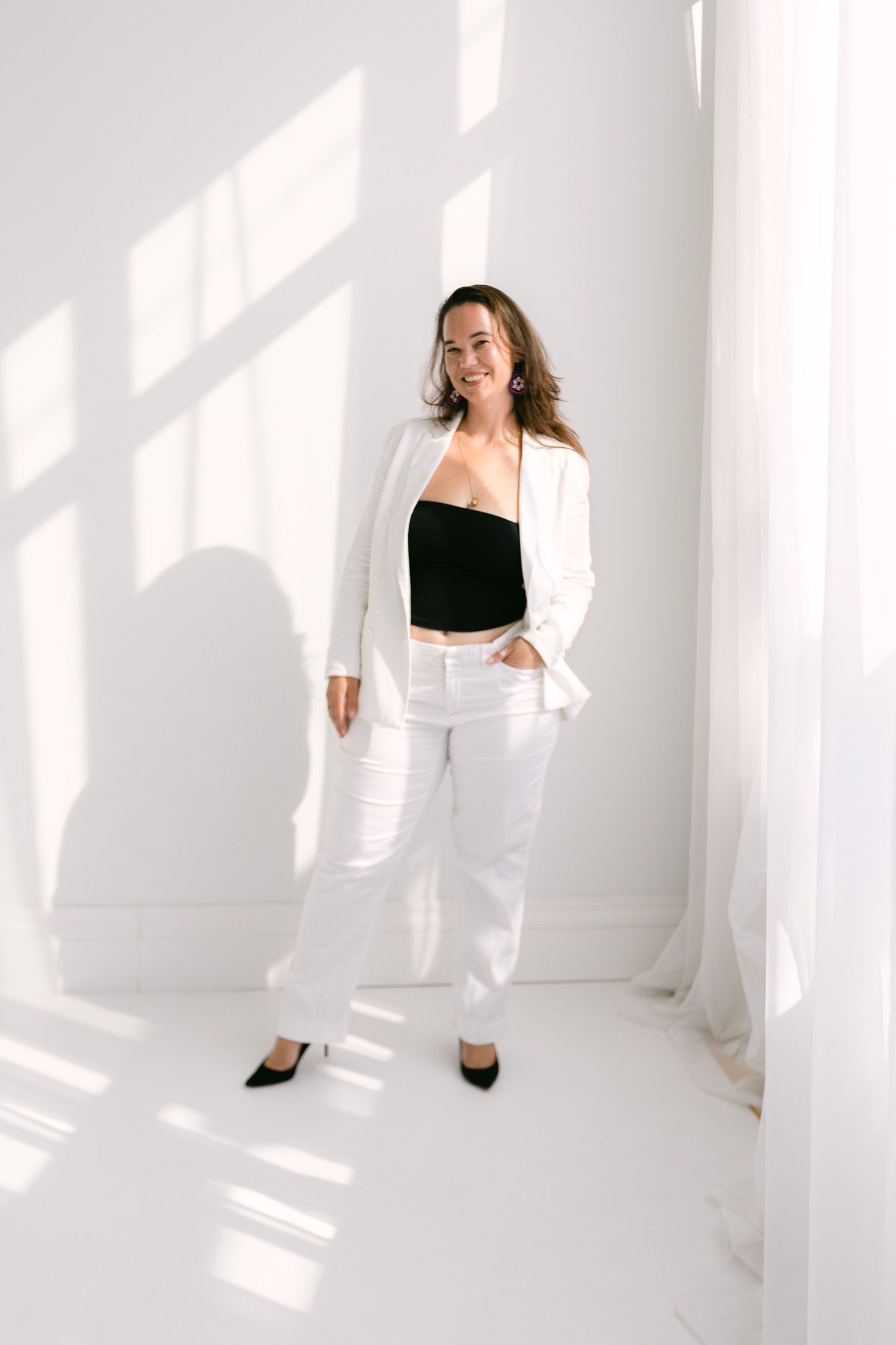 Woman in white blazer, white pants, and black crop top poses confidently in a bright white room with natural light.