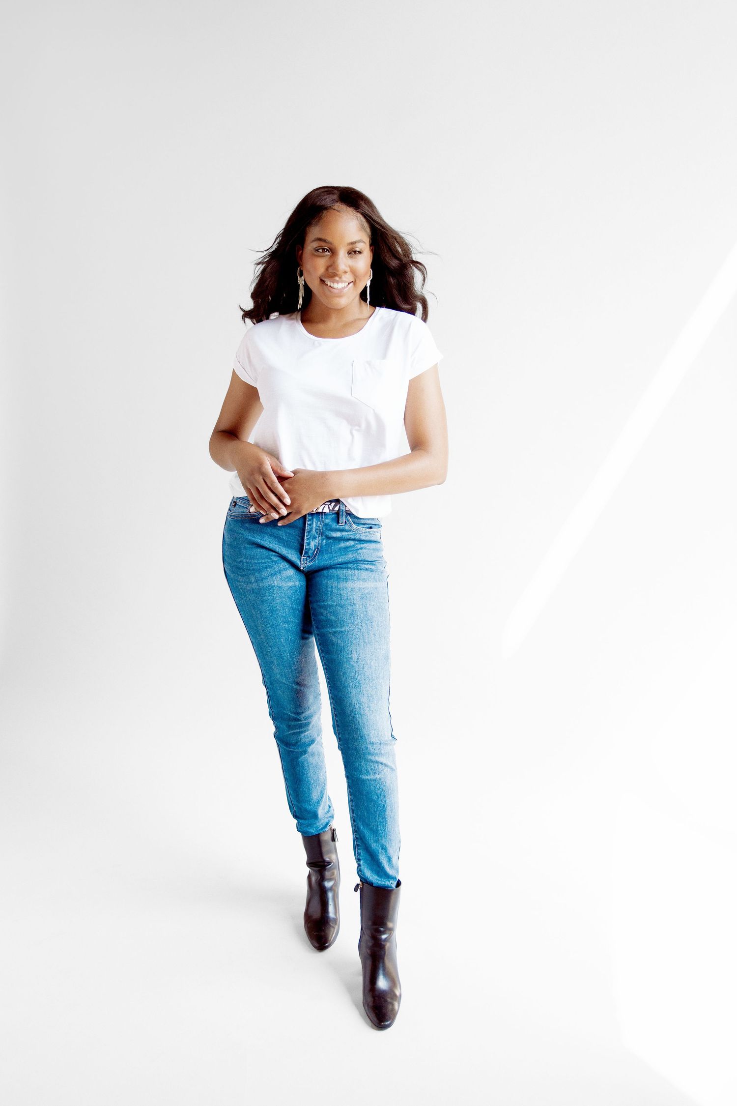 Smiling woman in white t-shirt and blue jeans with brown boots posing confidently against a white background.