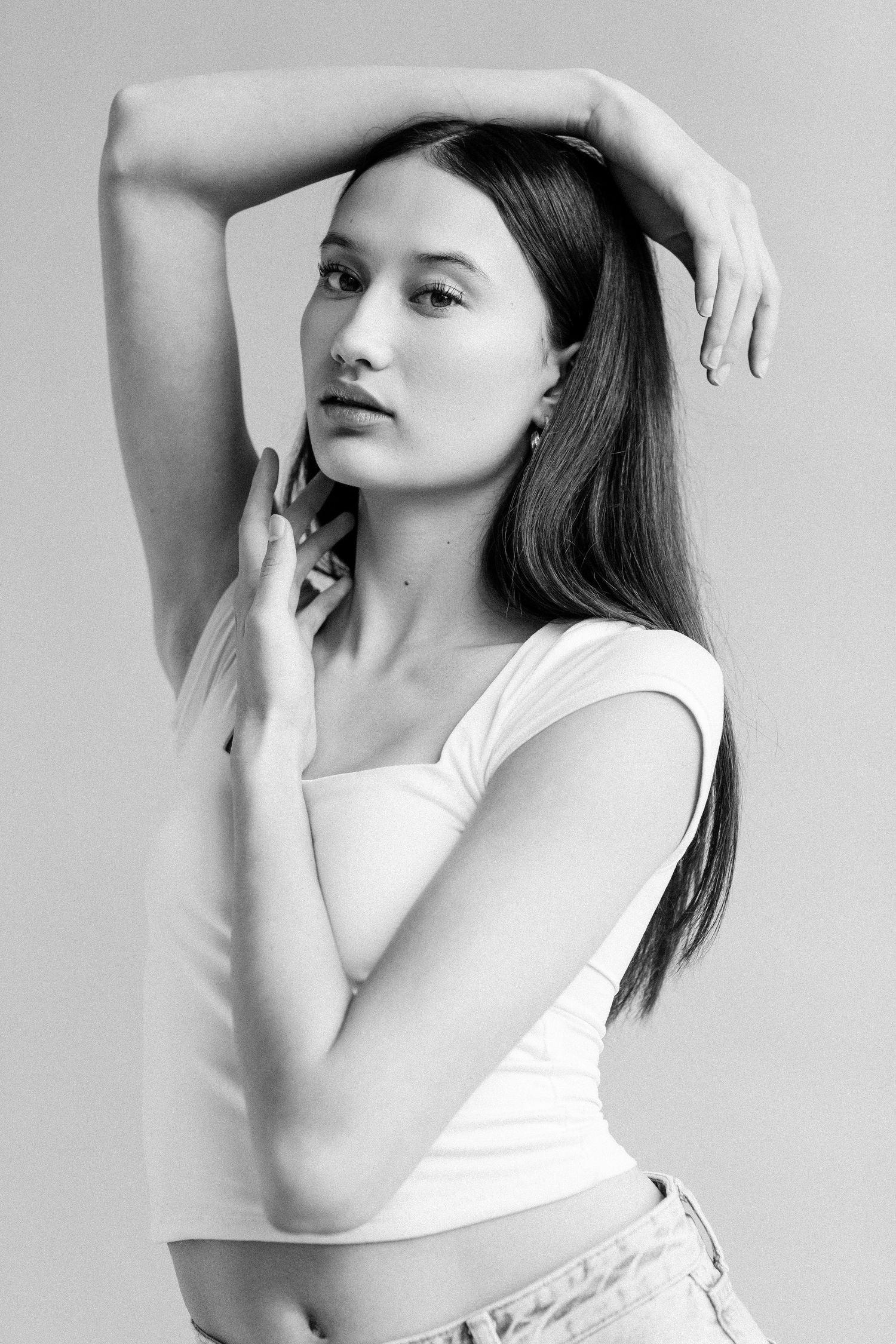 Black and white portrait of a young woman in a white crop top posing with arms raised, long hair flowing.