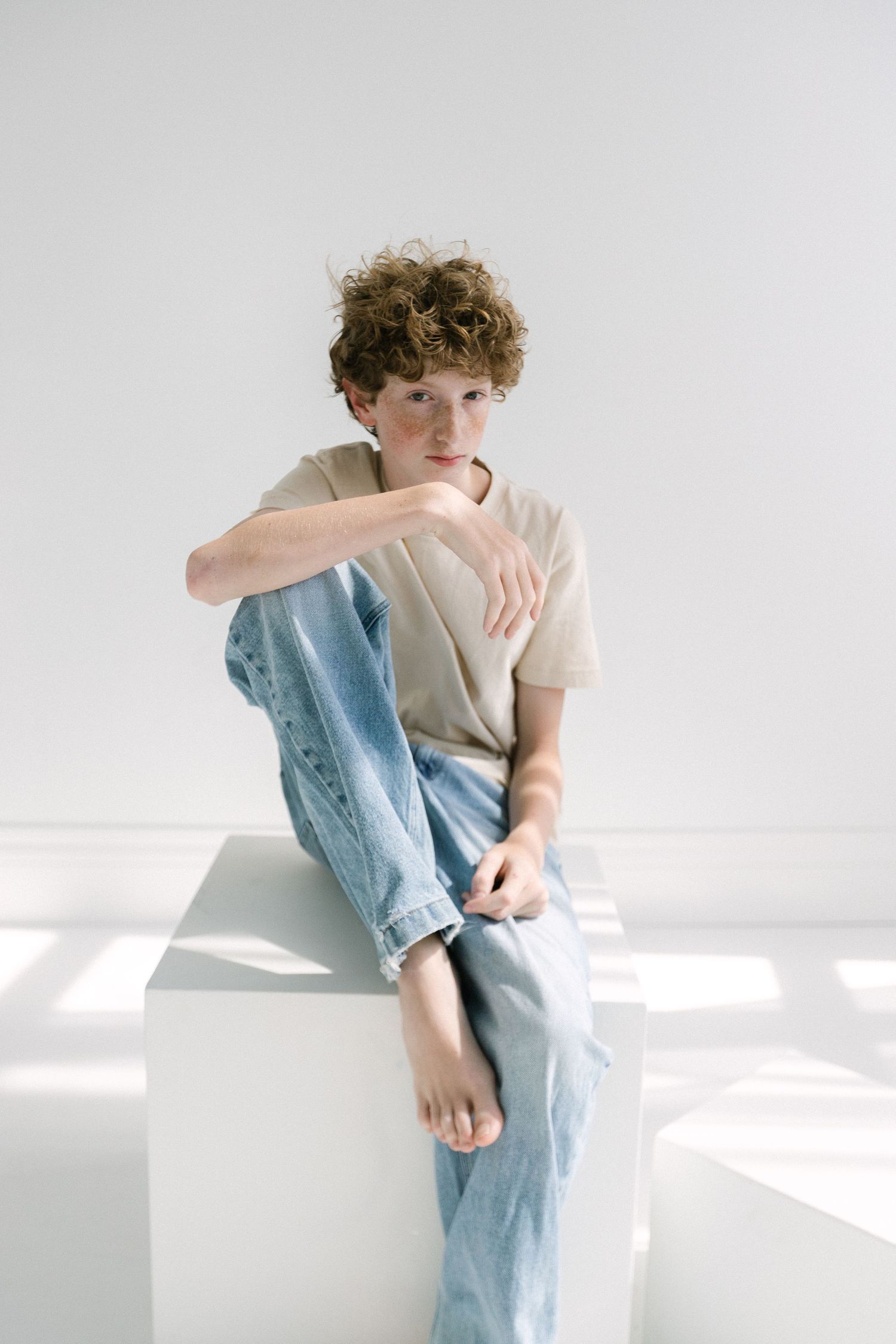 Young boy with curly hair sitting on white cube, wearing beige t-shirt and light blue jeans in minimalist studio.