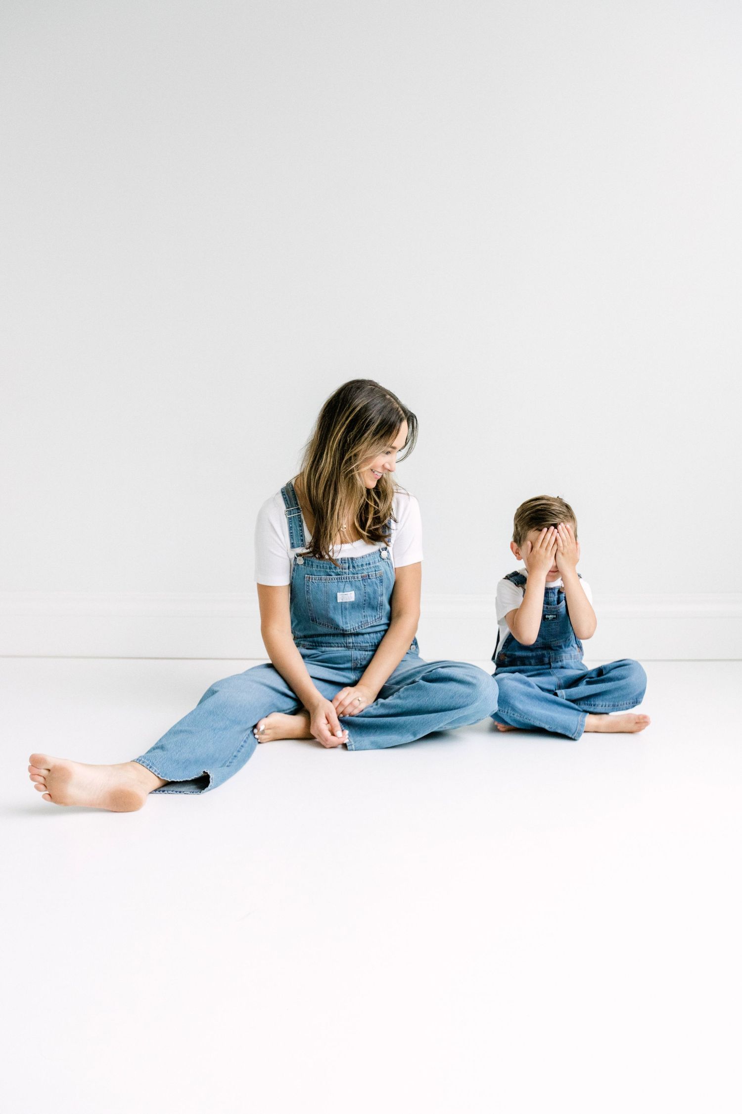 Mother and child sitting on white floor wearing matching denim overalls and white shirts in a bright studio setting.