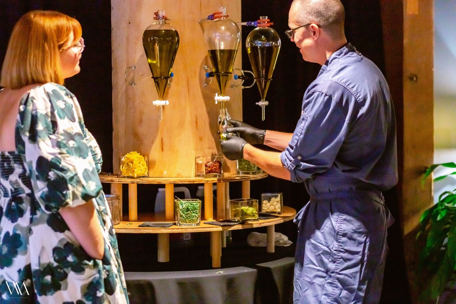 Man in blue uniform demonstrates botanical distillation equipment to woman at craft spirits exhibition.