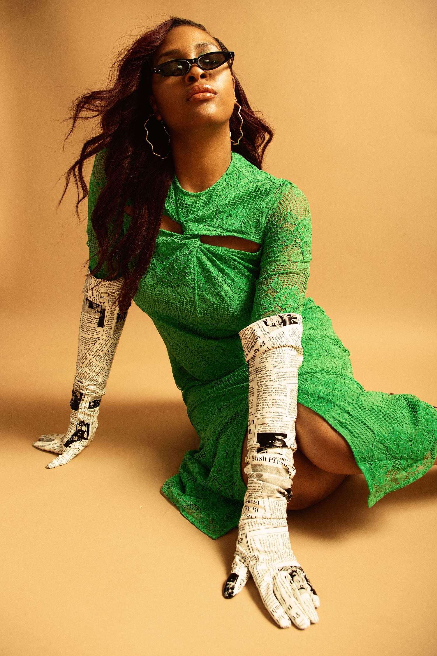 Woman in green lace dress and newspaper print gloves poses seated against beige background wearing dark sunglasses.