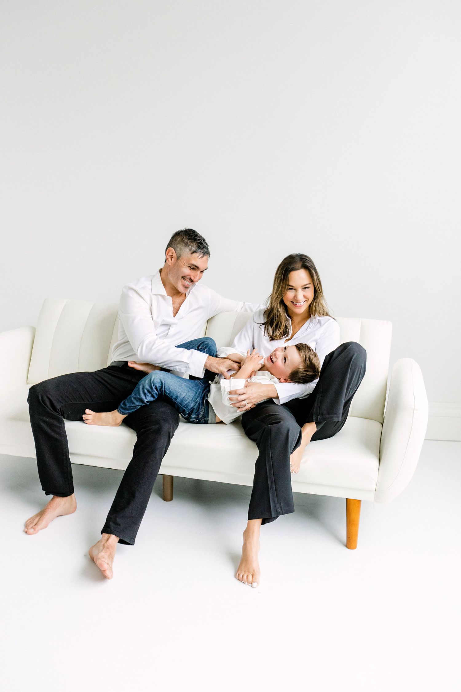 Happy family of three sitting on a white couch, parents in white shirts and black pants playing with their child.