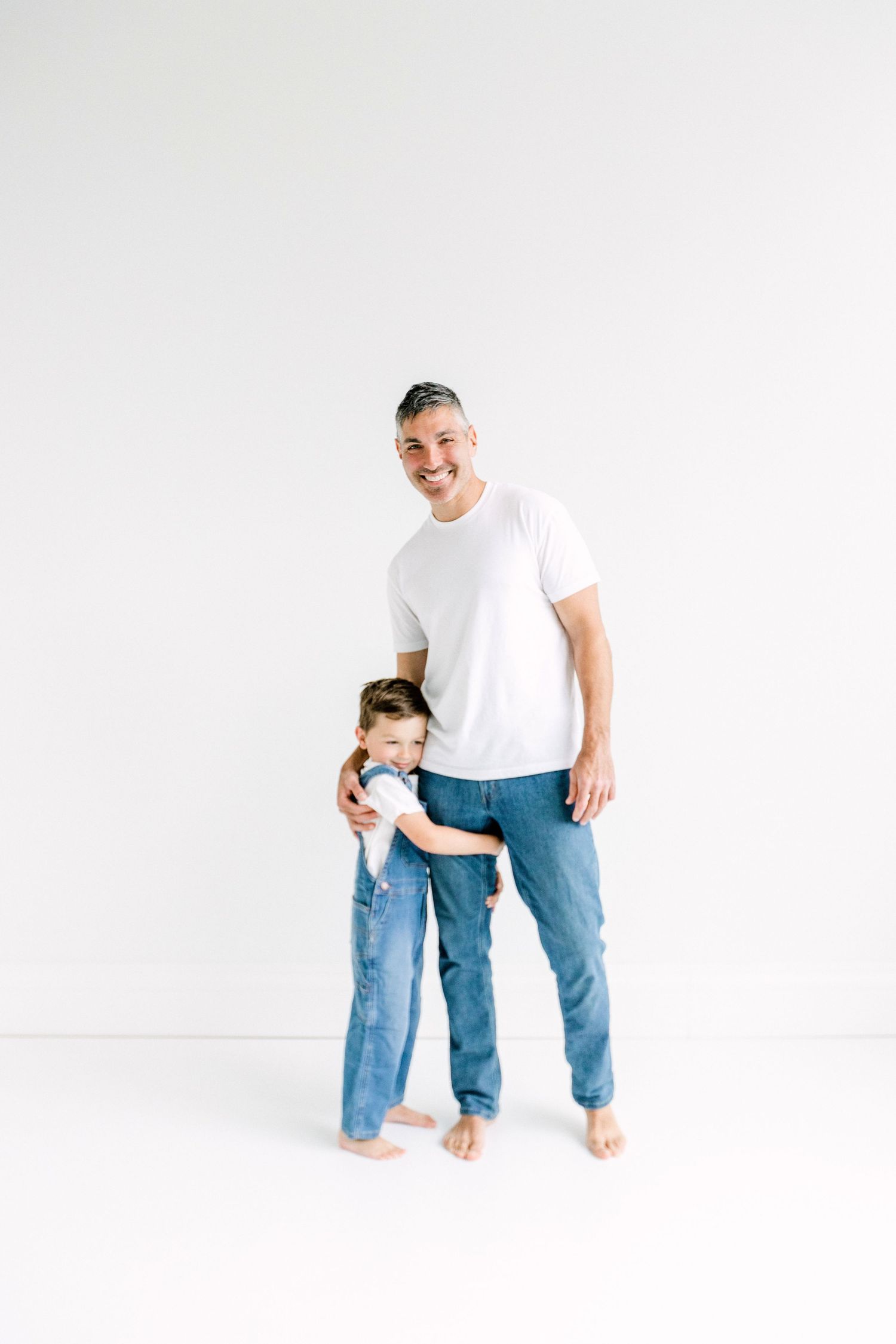 Smiling father in white t-shirt and jeans stands with young child hugging him against white background.