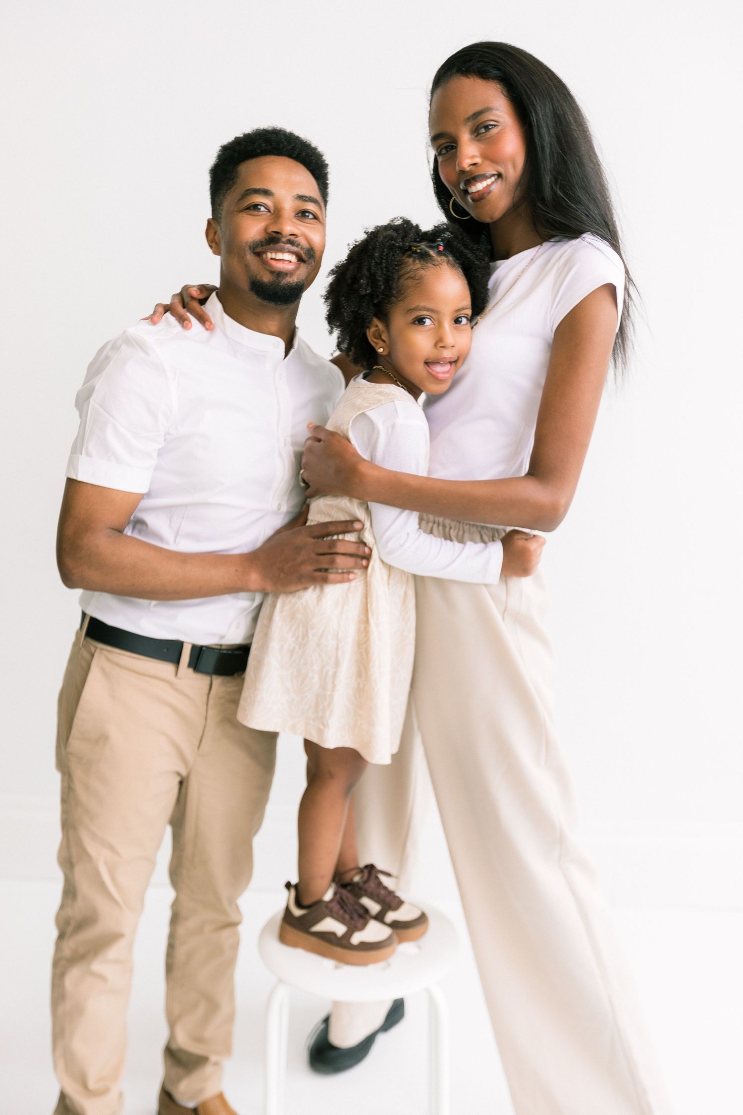 A happy Black family of three poses in matching white and beige outfits against a white background.
