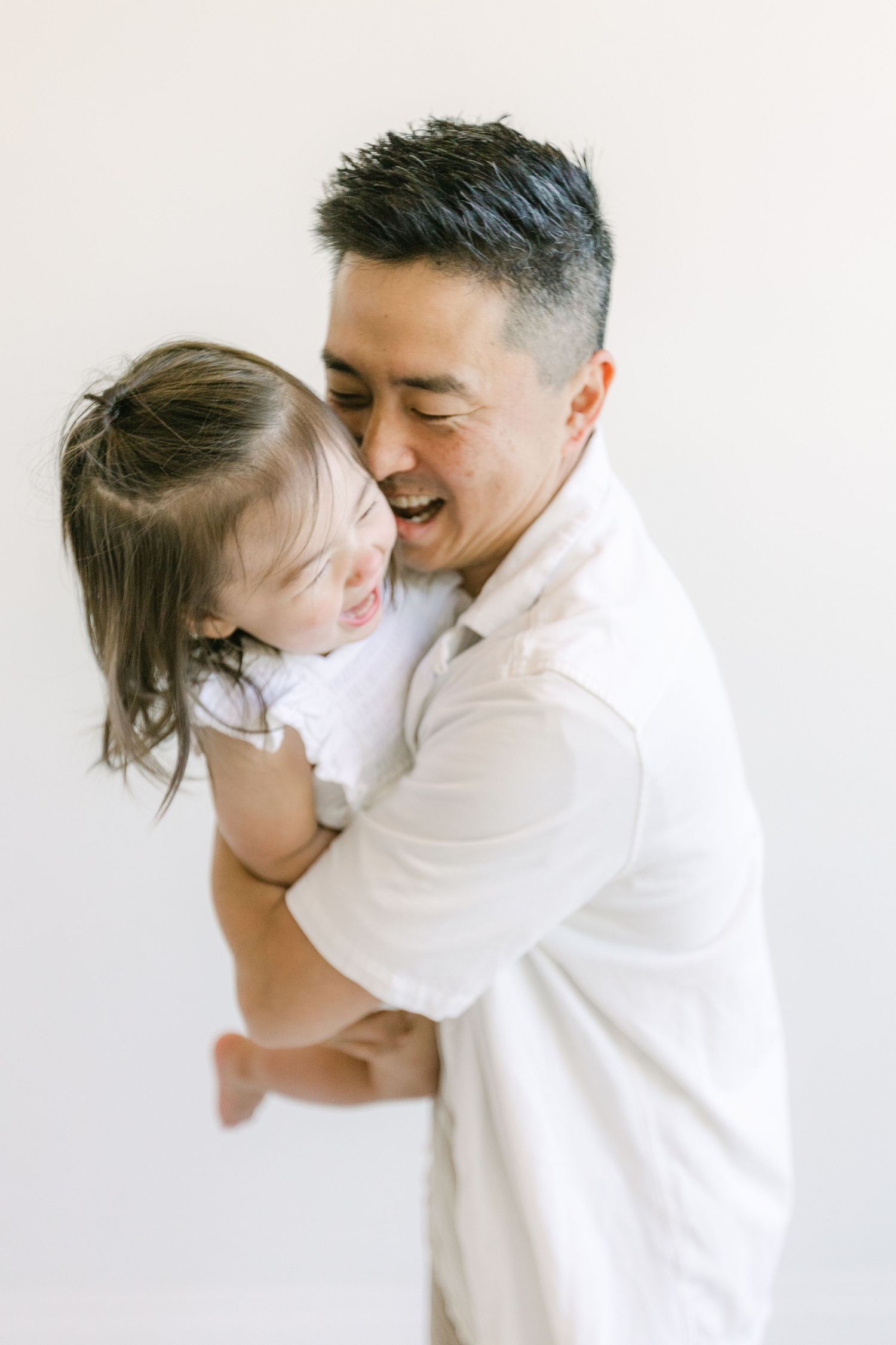 A smiling man in a white shirt joyfully holds a laughing young girl in a bright, minimalist setting.