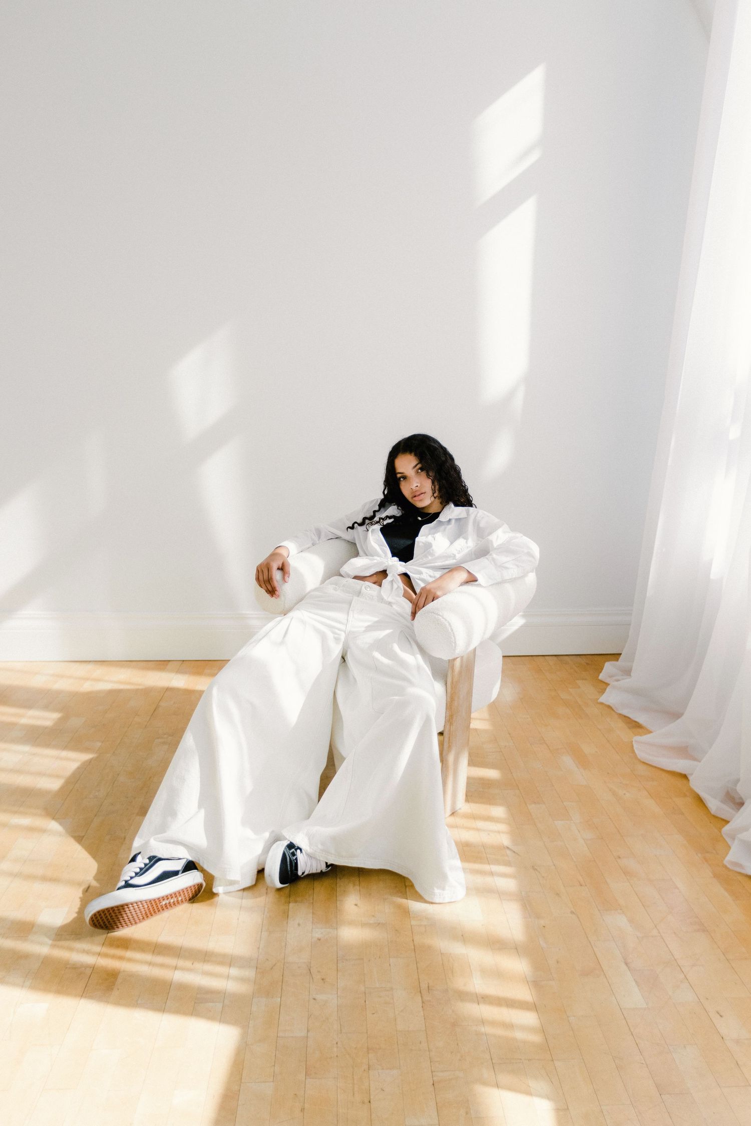 A young woman relaxes in a chair with sunlight streaming through white curtains.