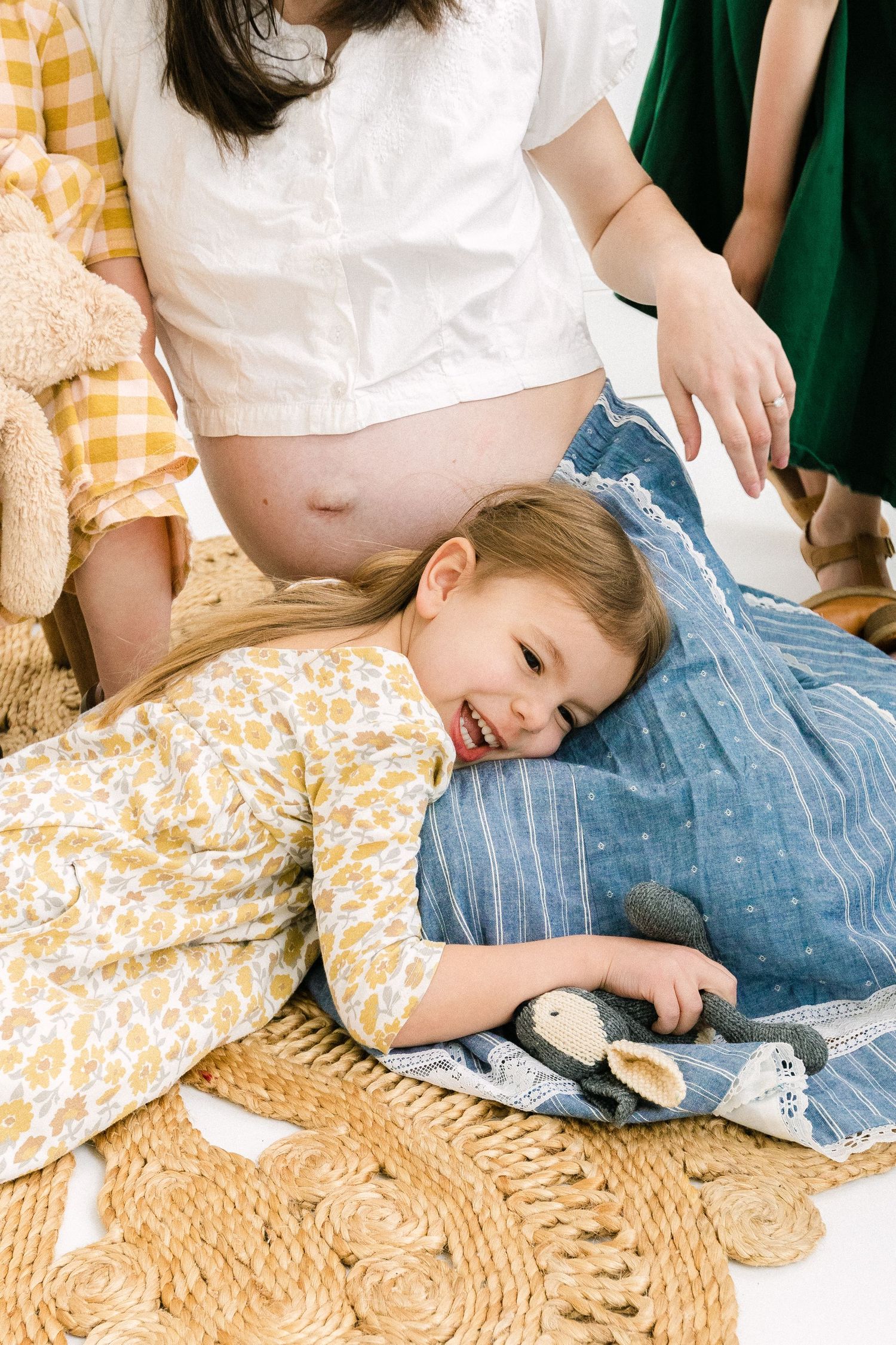 A smiling girl in a floral dress lays on a blue blanket near her pregnant mothers belly in a studio.