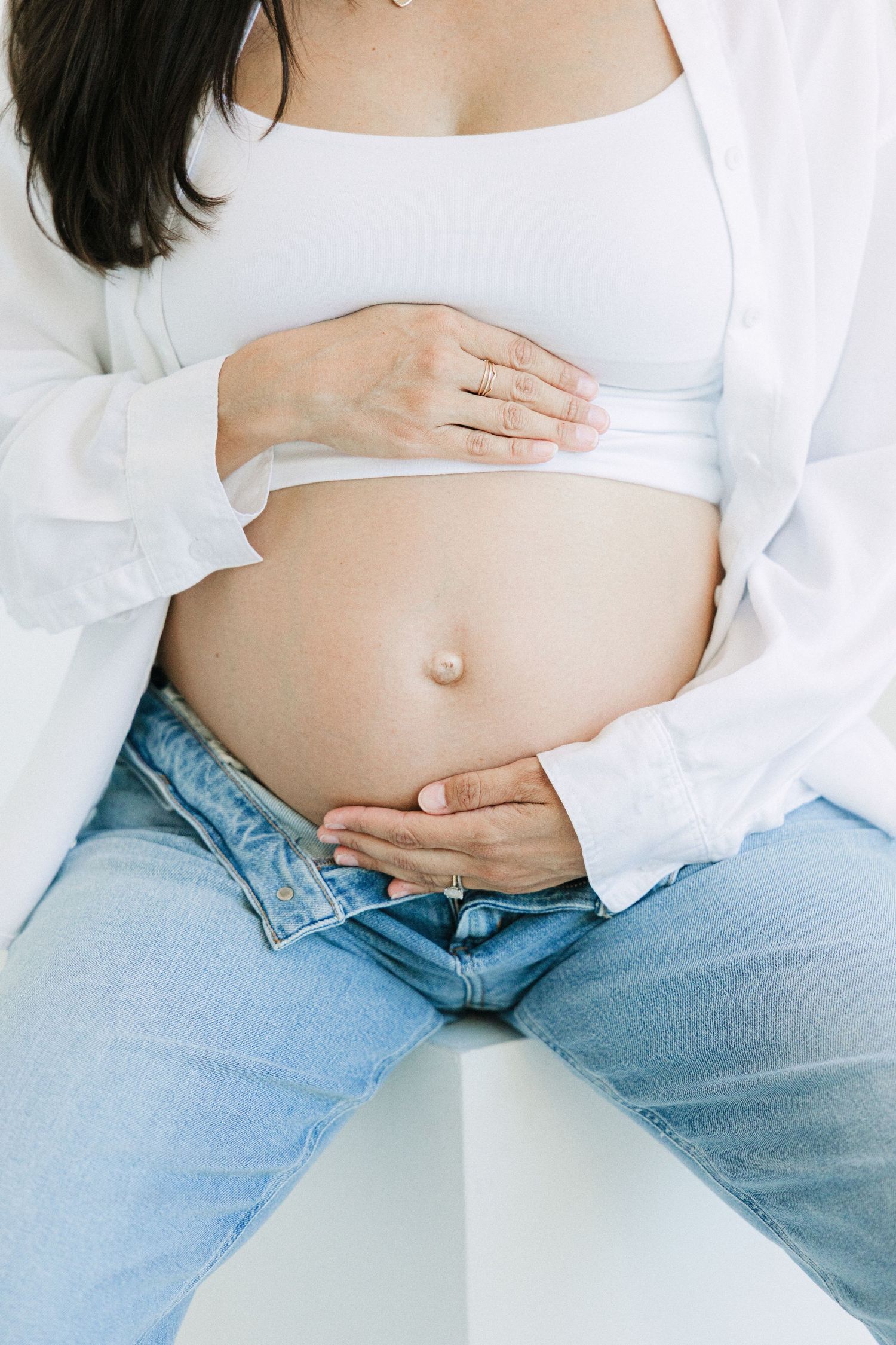 Pregnant woman in white crop top and open shirt cradling her bare baby bump with both hands, wearing blue jeans.