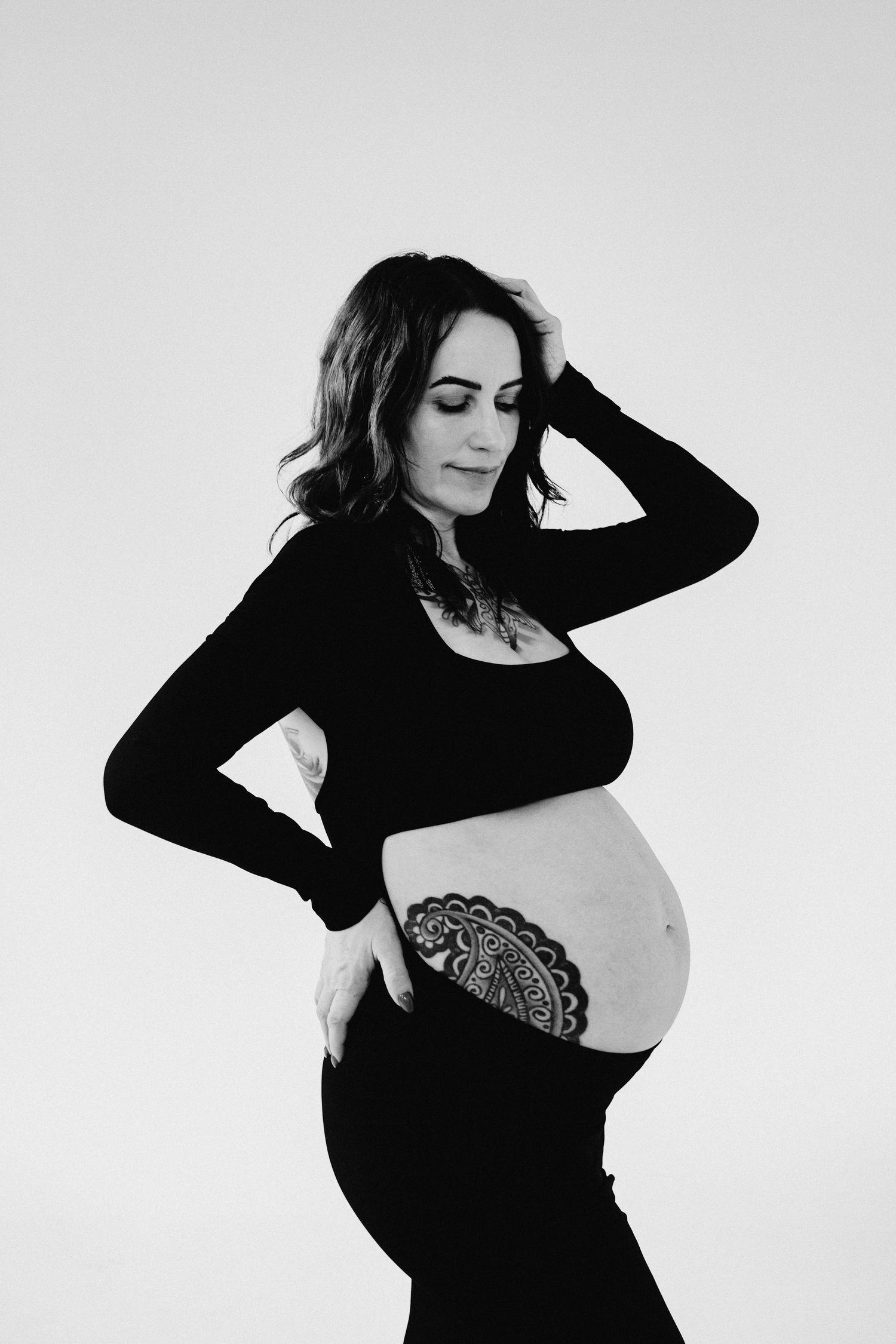 Black and white photo of a pregnant woman in black outfit showing her baby bump with henna art, posing gracefully.