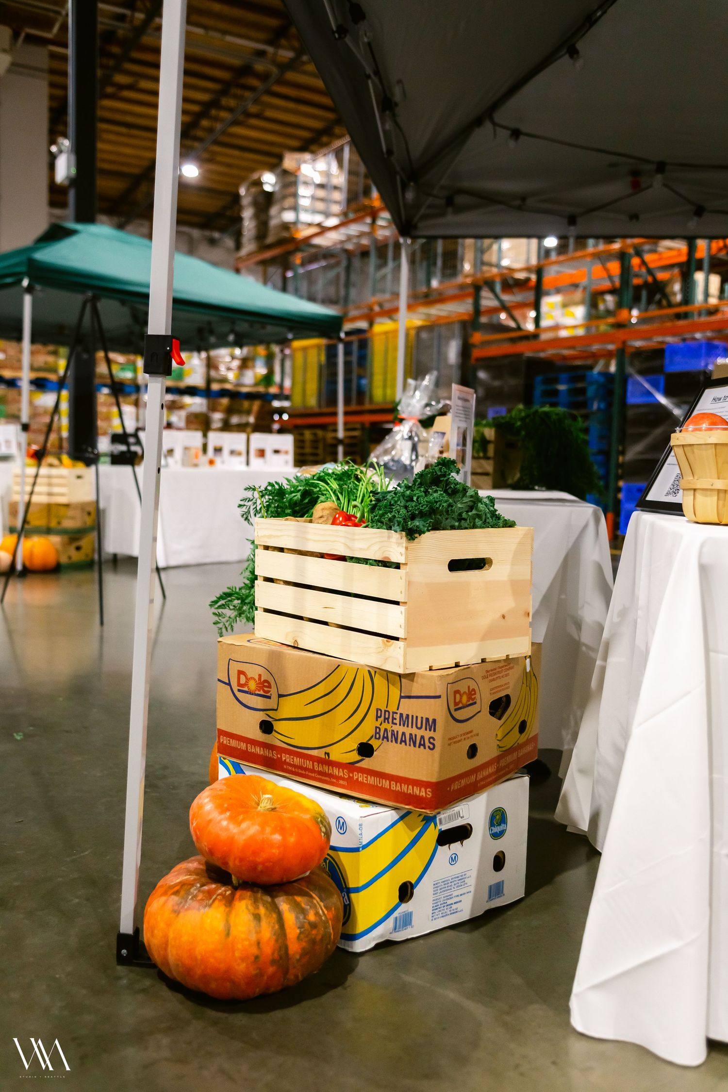Wooden crate of fresh vegetables stacked on Dole banana box with orange pumpkins on warehouse floor.
