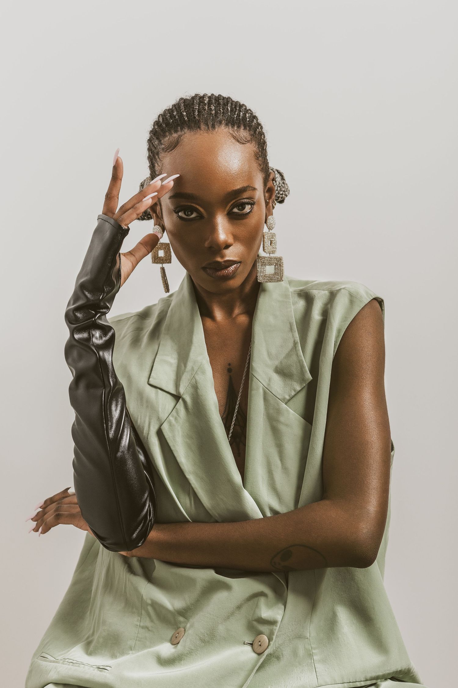 Stylish Black woman in sage green sleeveless blazer with leather glove and geometric earrings posing confidently.