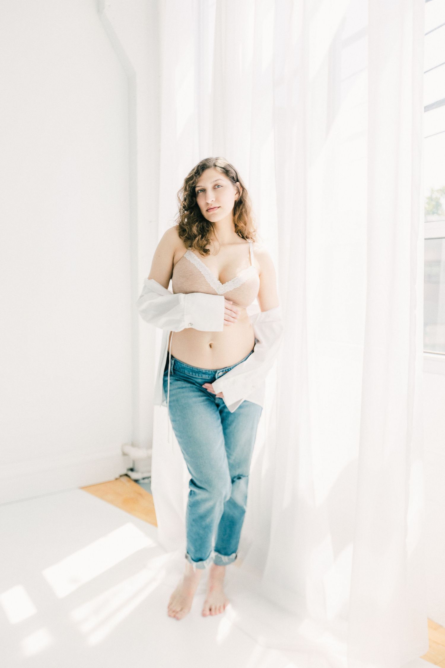 Pregnant woman in beige bra, open white shirt, and jeans standing by sheer curtains in bright white room.