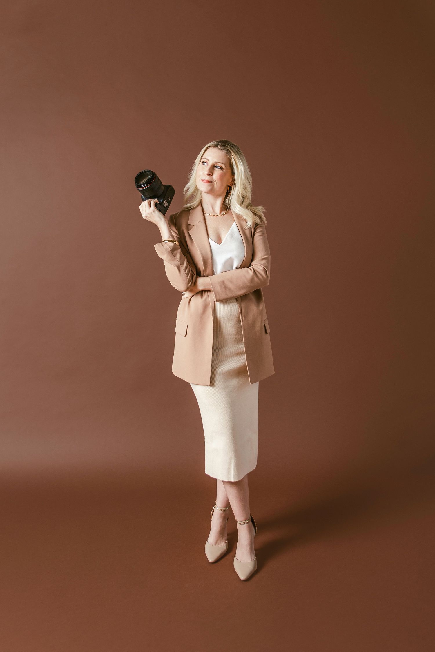 Blonde woman in beige blazer and white dress holding a camera on brown background.