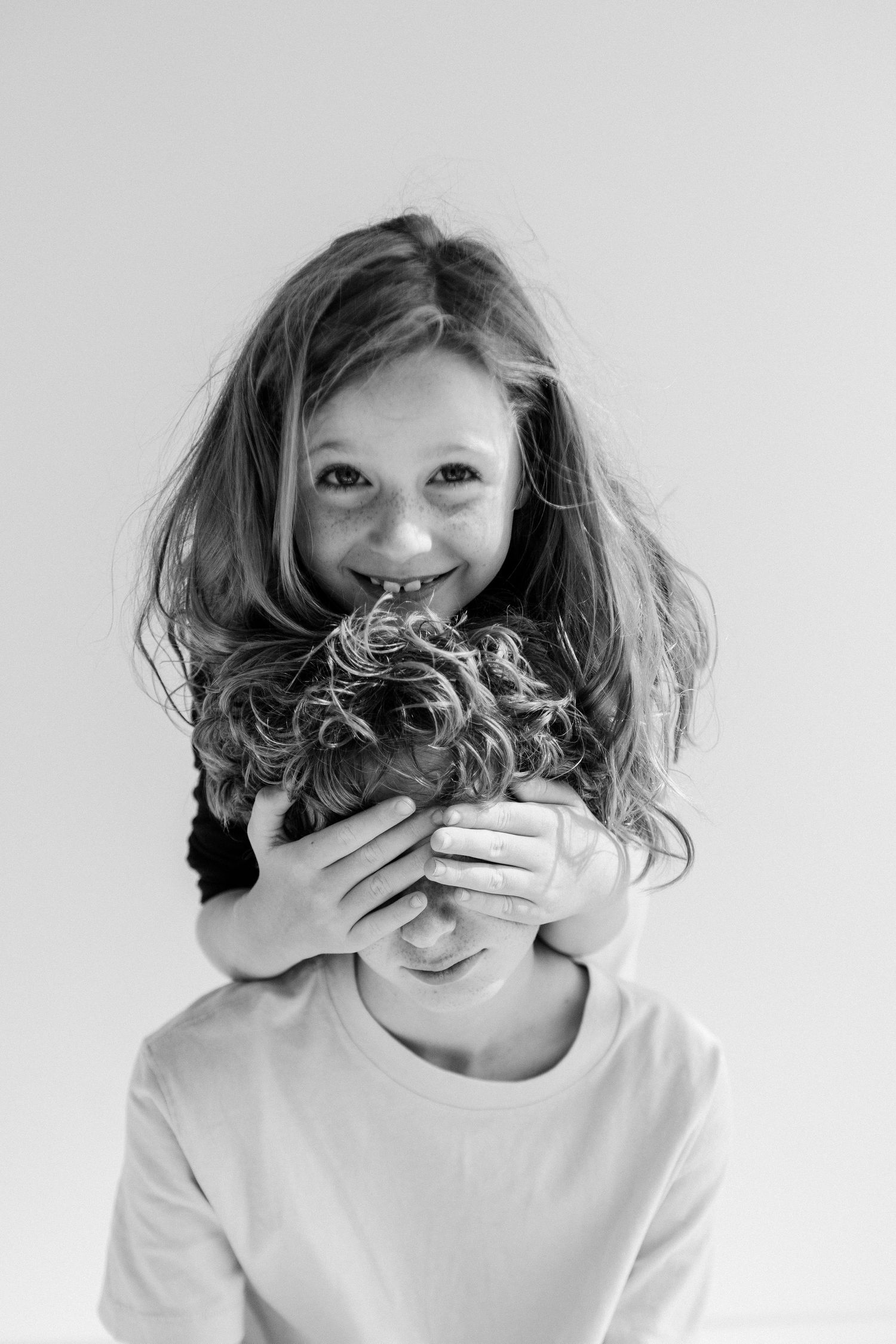Black and white photo of a smiling girl covering a younger child's eyes with her hands in a playful pose.