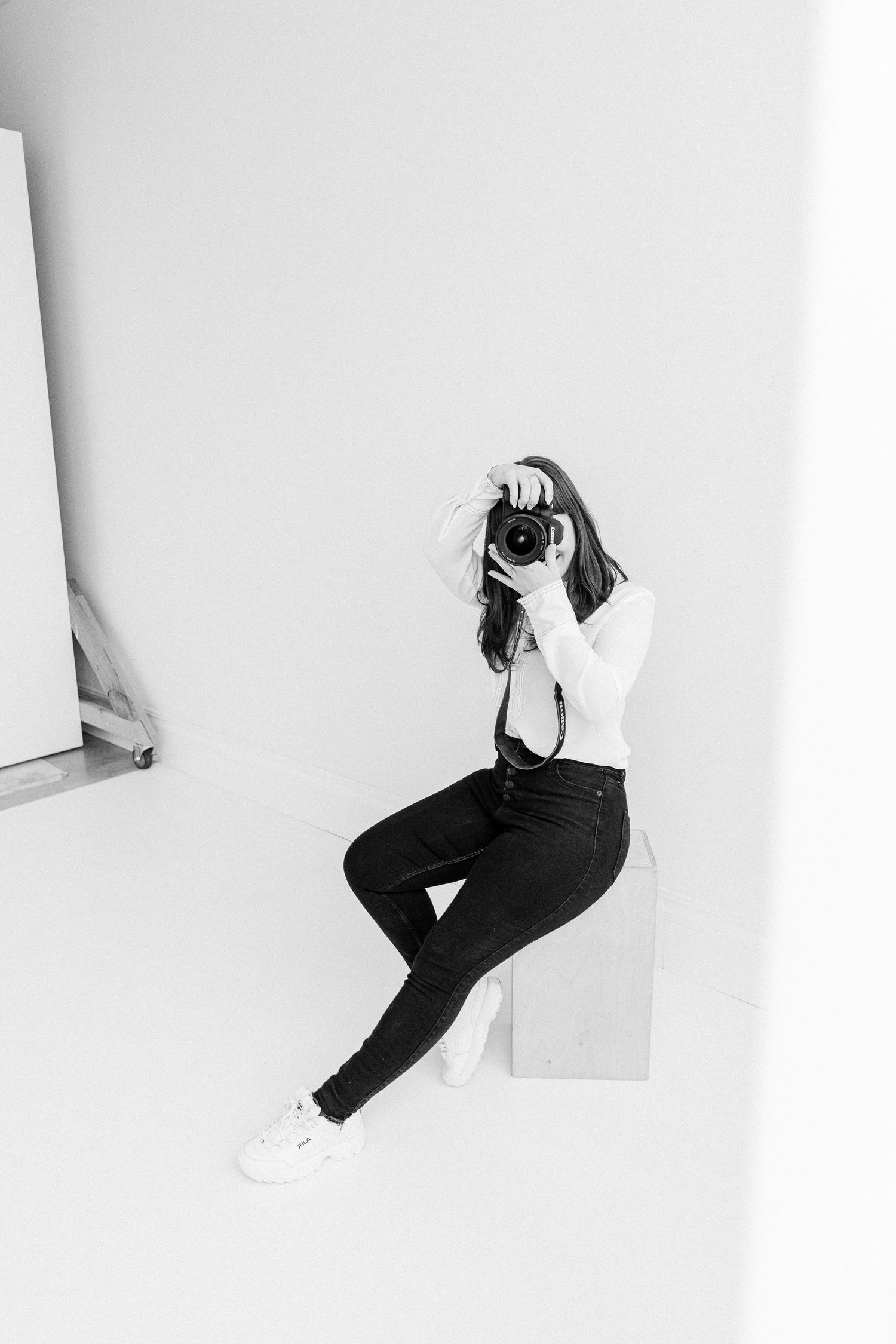 Female photographer sitting on a cube in a white studio, holding a camera up to her face, black and white.