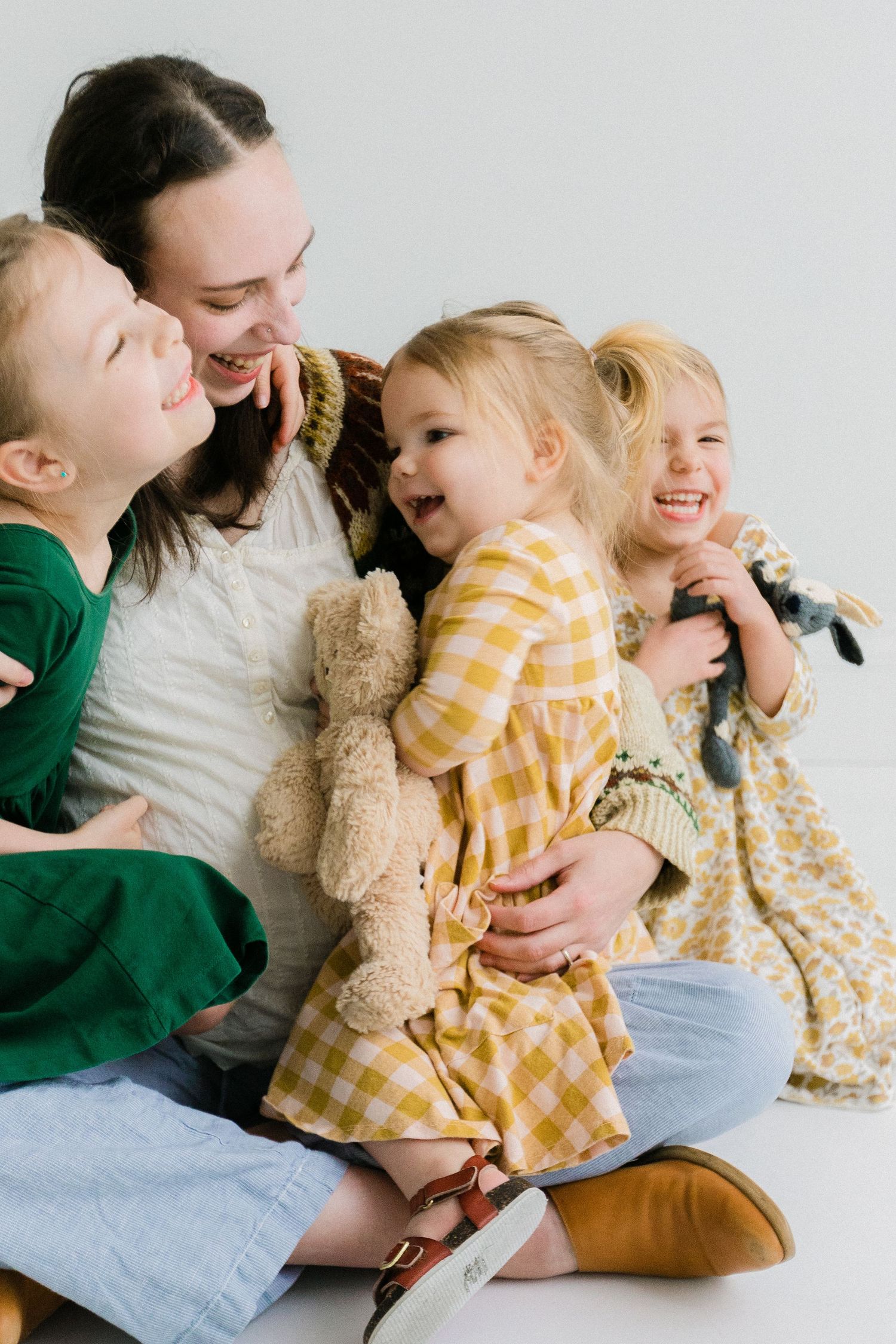 A joyful mother sits on the floor laughing with her three young children holding stuffed animals.