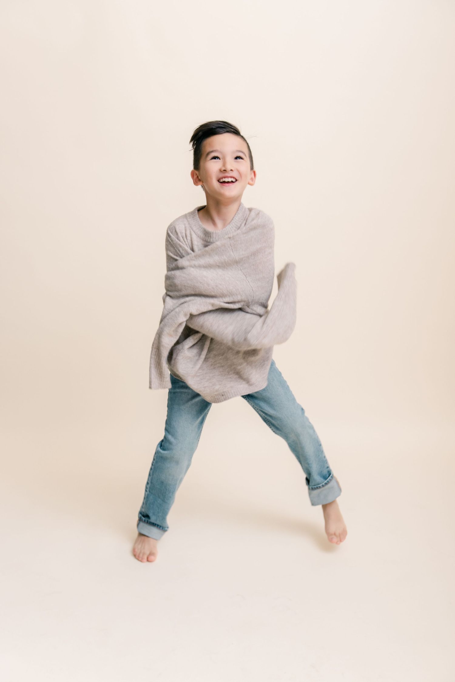 A happy barefoot boy laughs while wrapped in an oversized gray sweater in a studio.