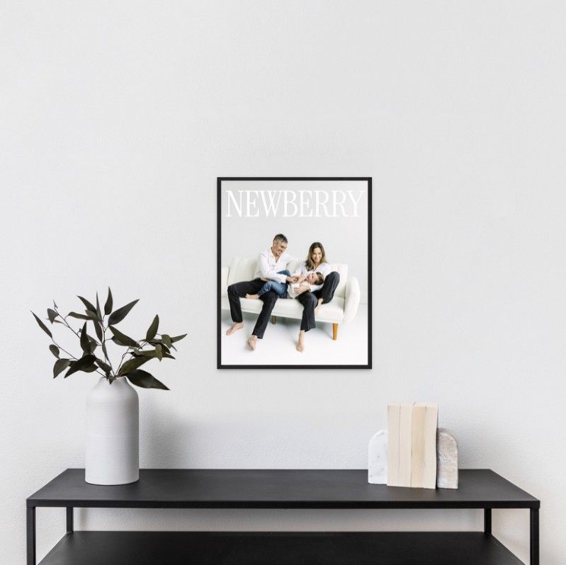 Framed family portrait print with NEWBERRY text displayed on a black console table with a white vase and plant.