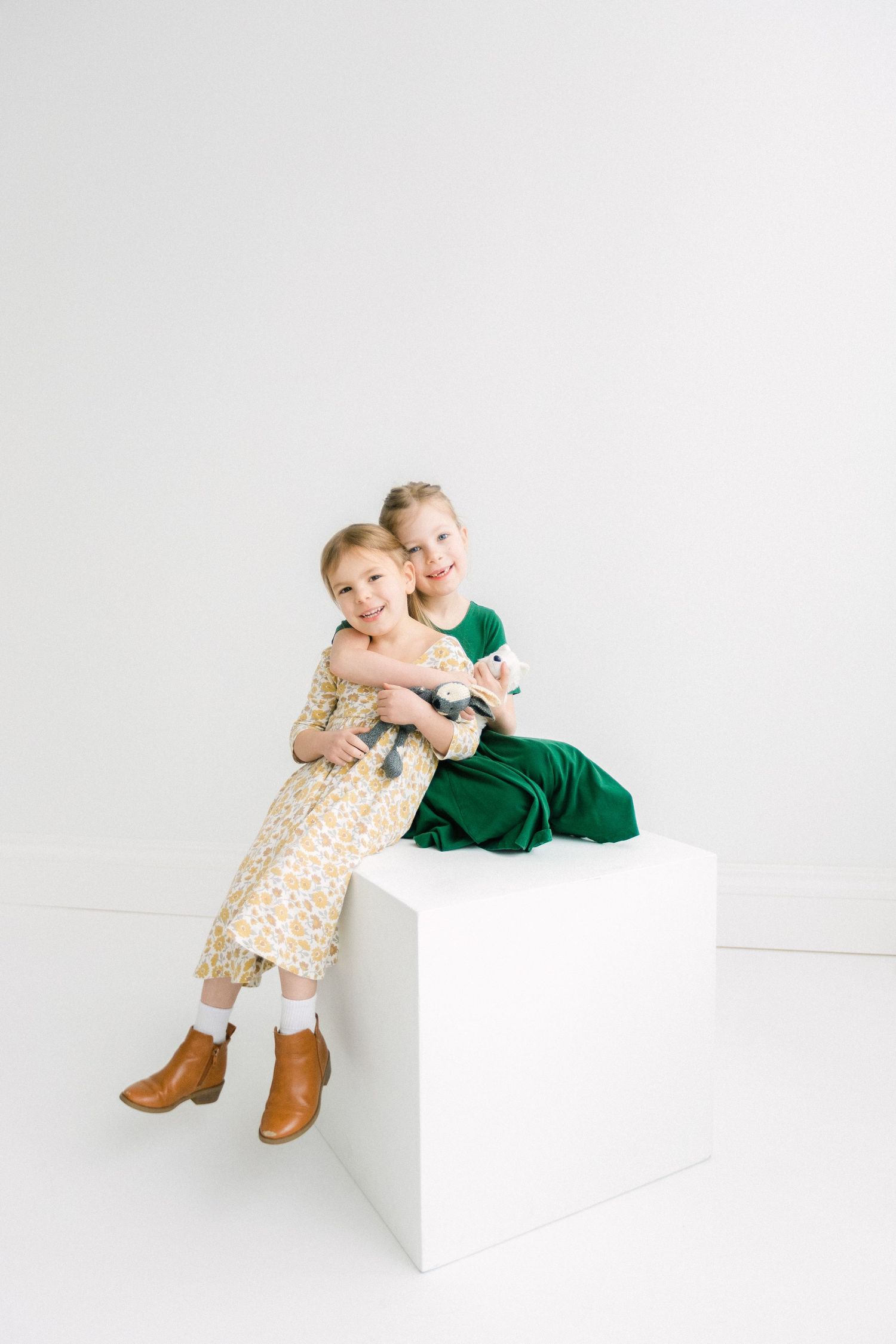 Two young girls in dresses, one floral and one green, hugging and smiling on a white cube in a bright studio.