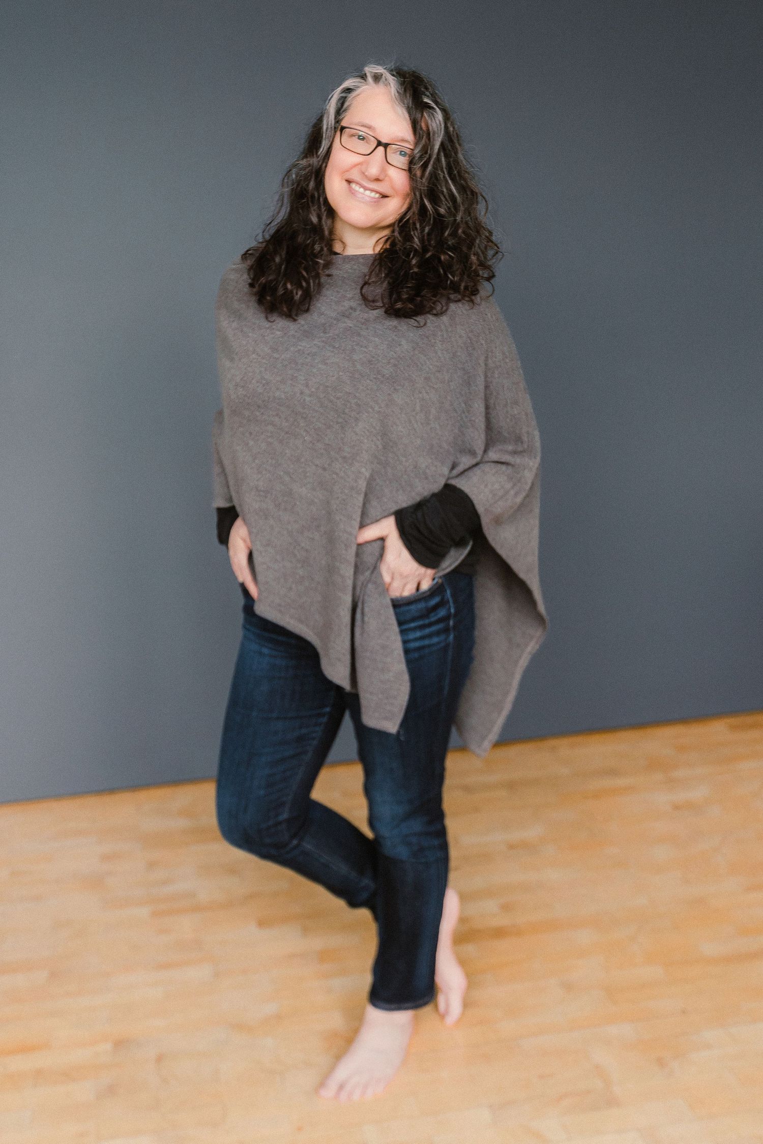 Smiling woman with curly hair and glasses wearing a gray poncho and jeans, posing barefoot in a studio.