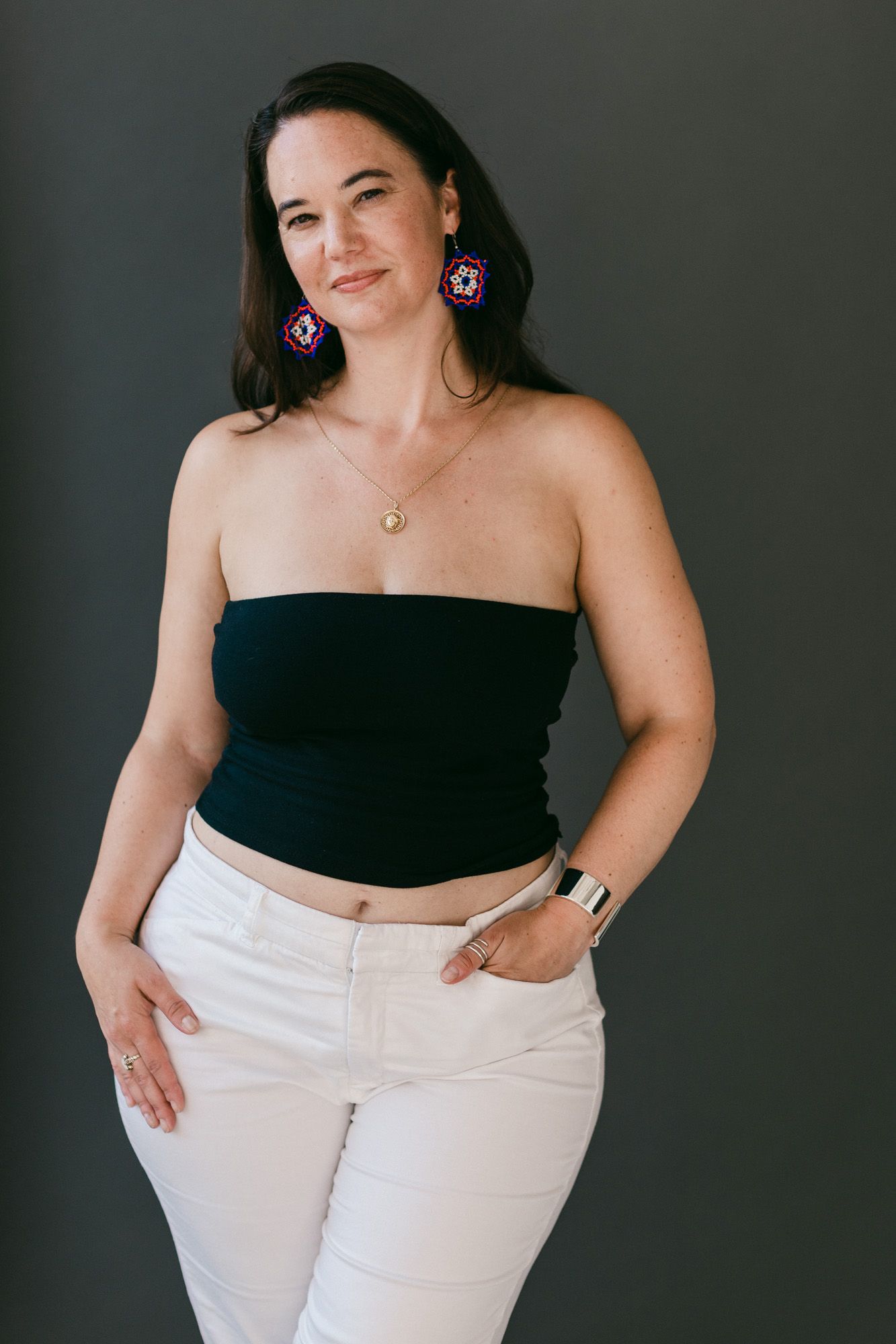 Woman in black tube top and white jeans wearing colorful earrings and gold necklace posing against gray background.