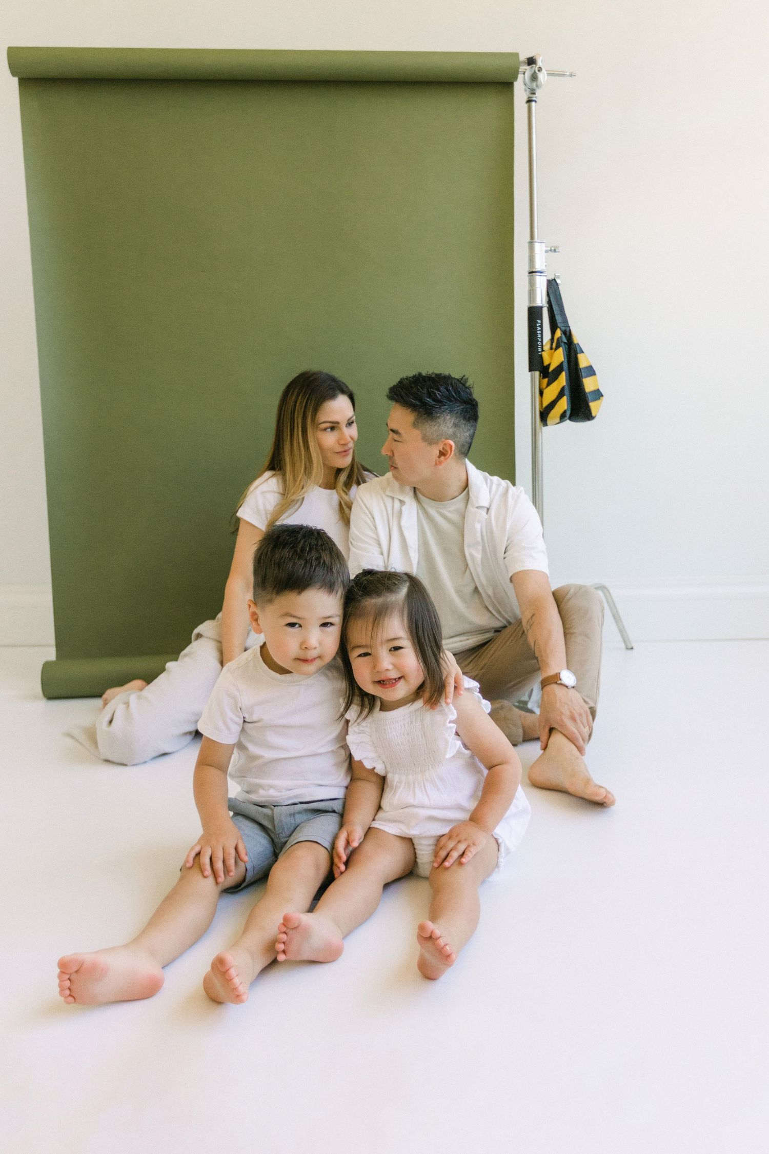 Asian family of four posing in photo studio with olive green backdrop, parents sitting behind two young children.