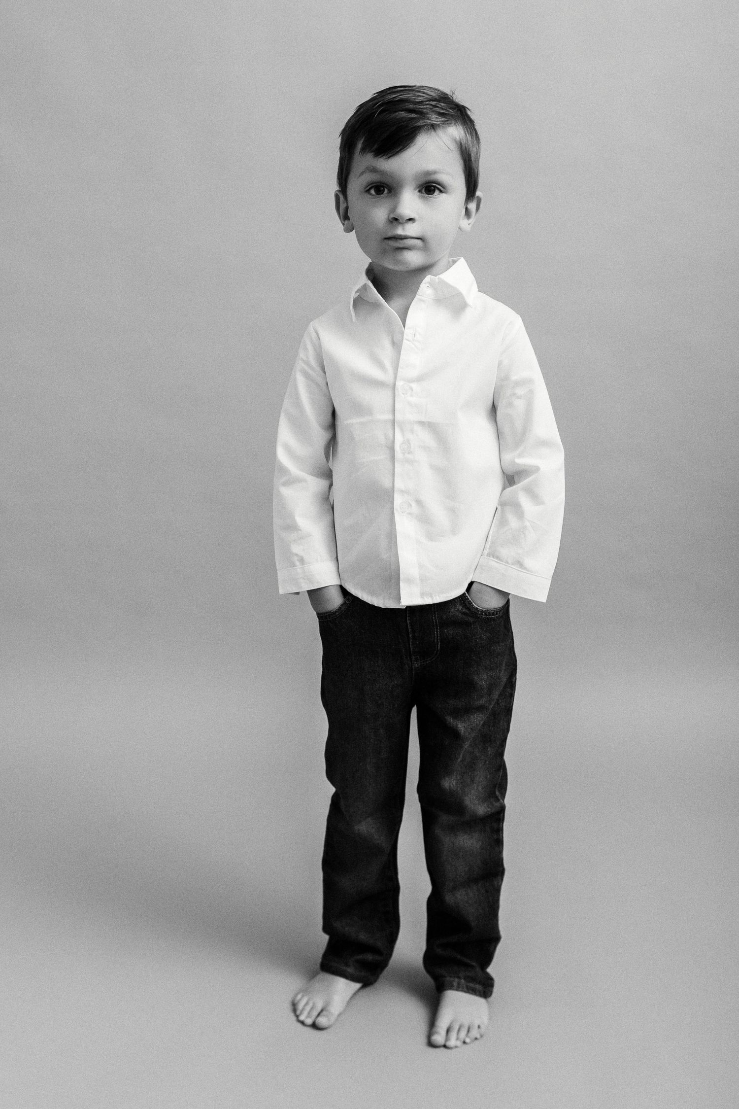 Black and white portrait of a young boy in white shirt and jeans, standing barefoot against a neutral background.