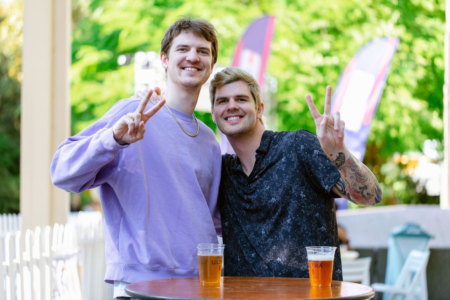 Two smiling young men making peace signs at outdoor bar table with glasses of beer.