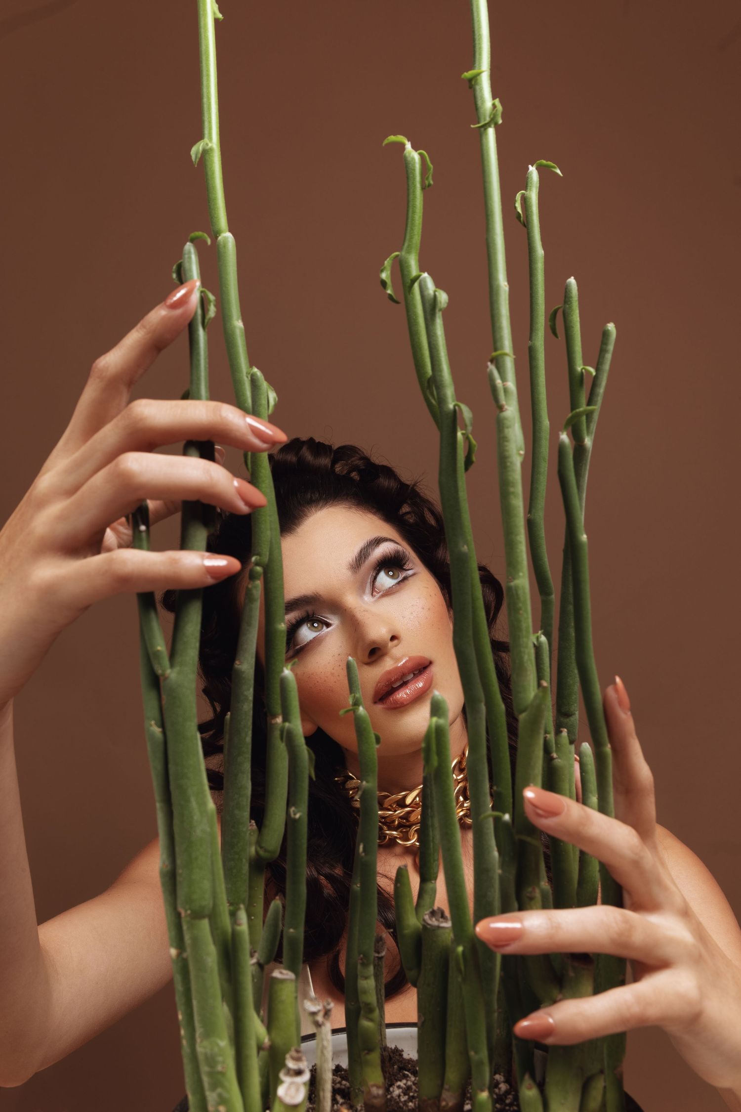 Young woman with gold jewelry peers through tall green succulent stems against a brown background.