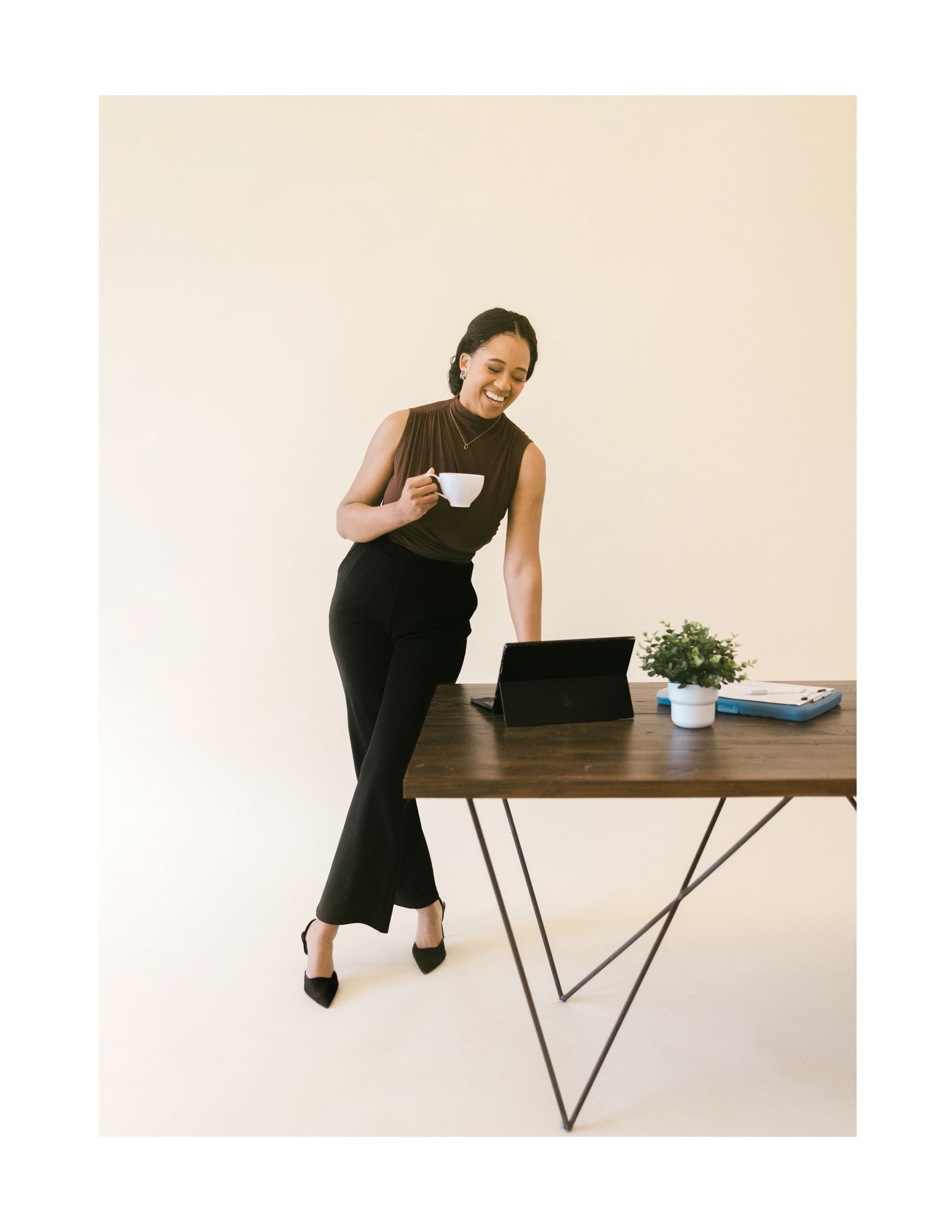 A professional woman in black wide-leg pants holds a coffee cup while leaning against a modern desk with a tablet.