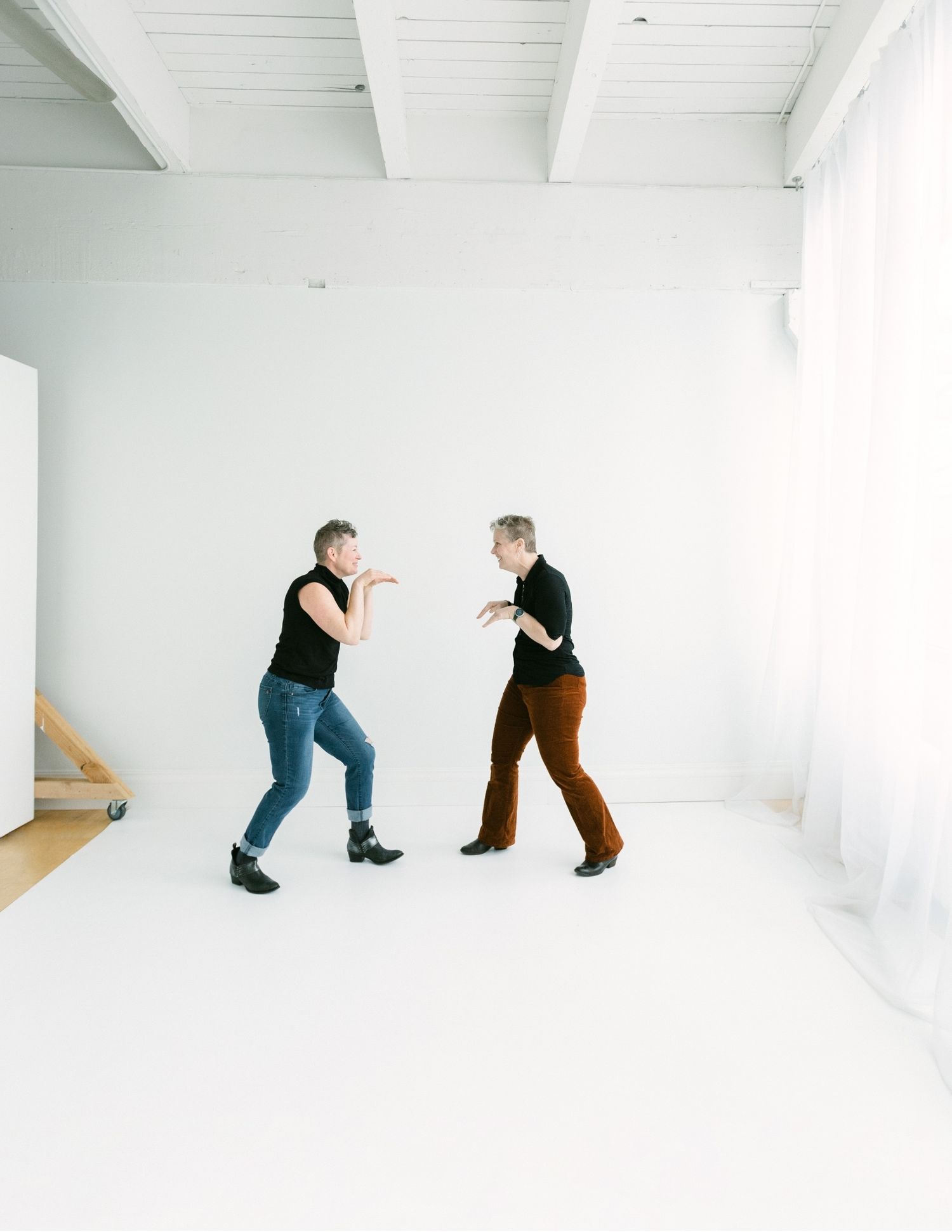 Two women in black tops playfully face each other in a fun dance pose in a white studio.