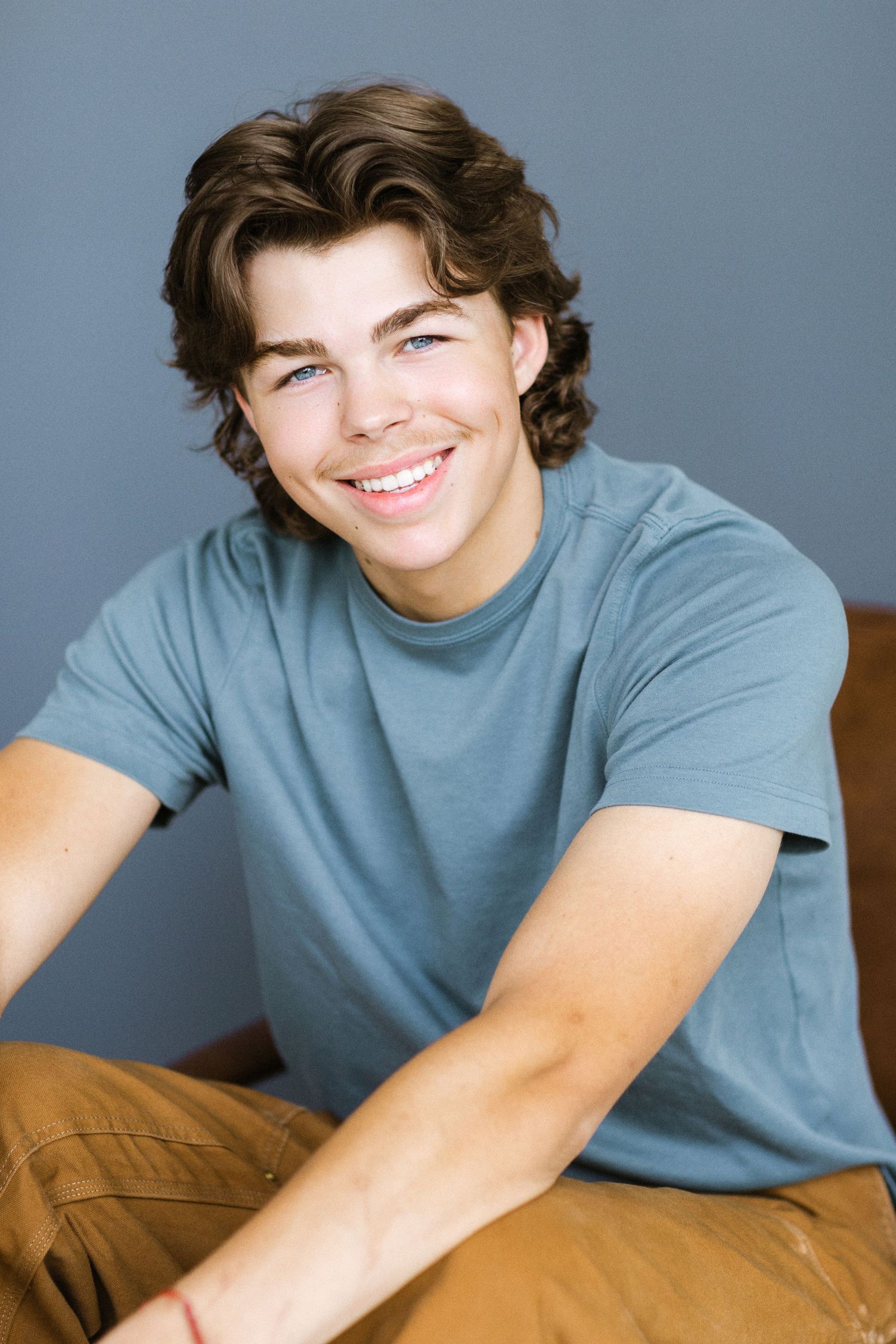 Smiling young man with brown hair wearing a teal t-shirt and tan pants, seated against a gray background.