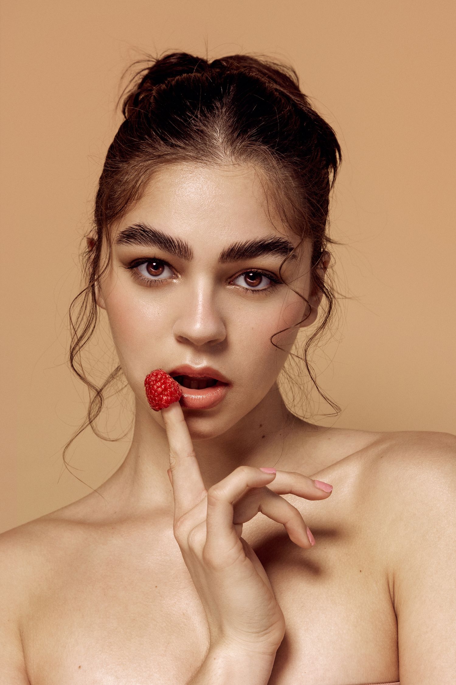 Beauty portrait of a young woman with bold brows holding a fresh raspberry to her lips against beige background.